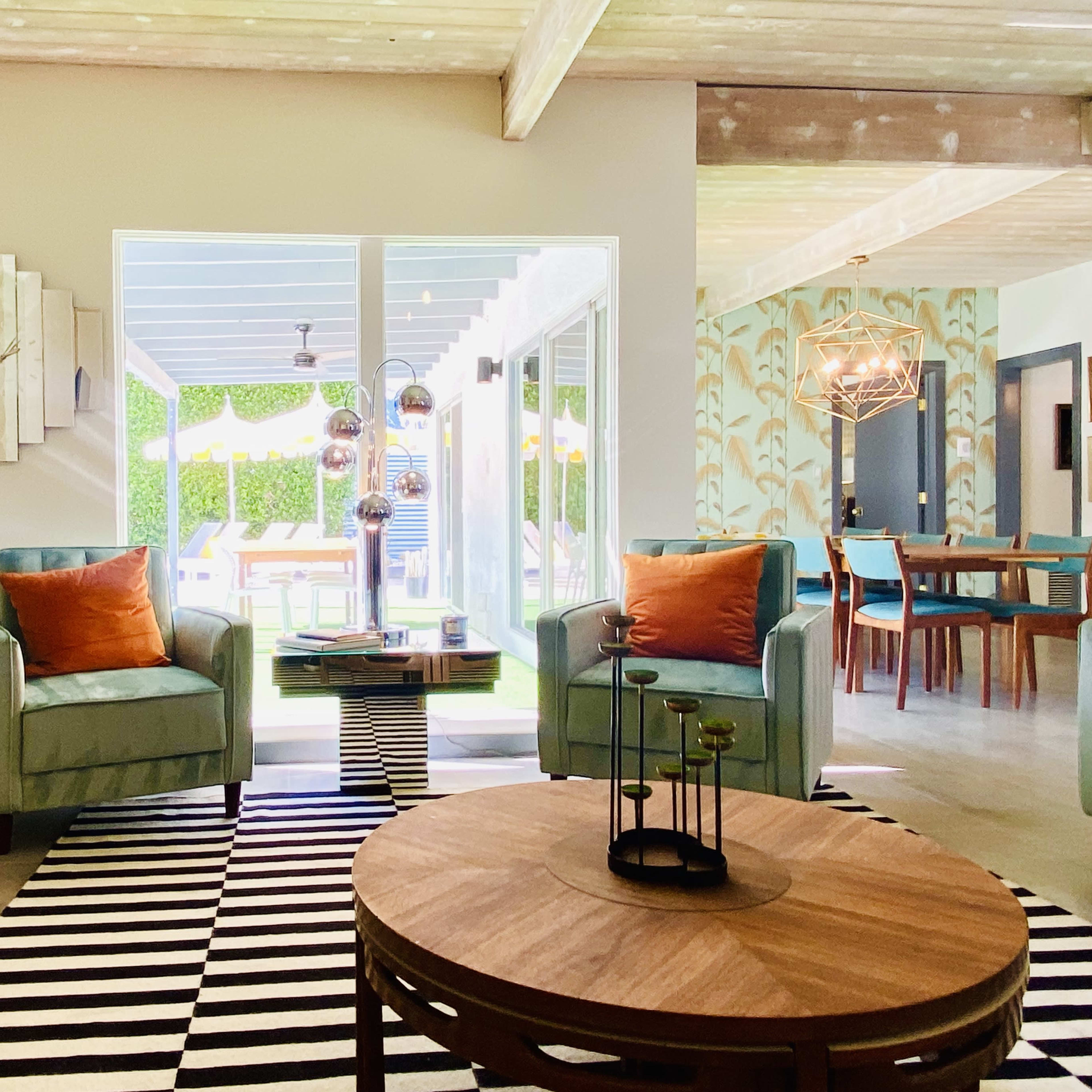 A modern living room featuring two teal armchairs with orange pillows, a round wooden coffee table, and large windows overlooking an outdoor area with patio furniture.