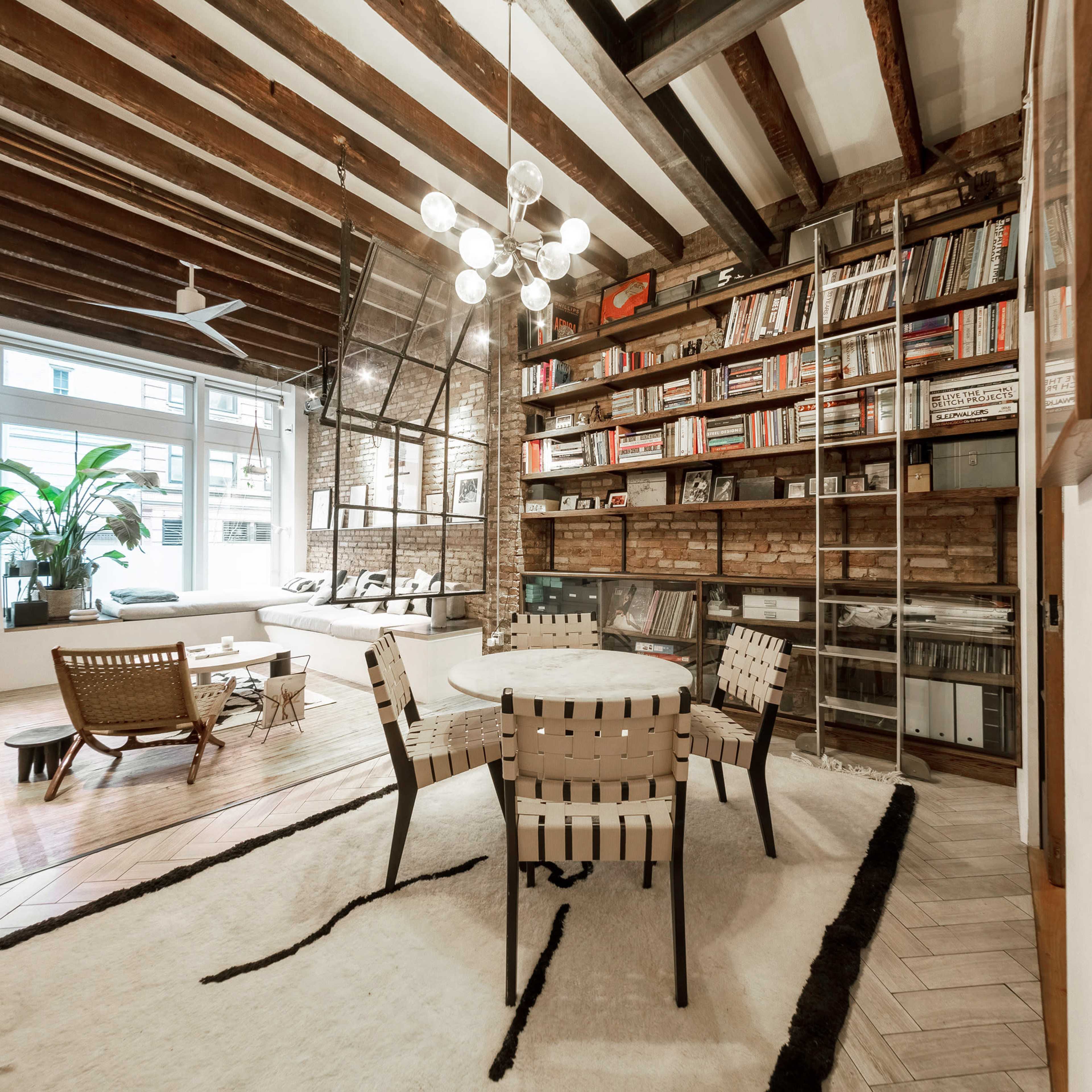 A modern living space features a circular table surrounded by four chairs, with a large bookshelf and a window showcasing natural light.