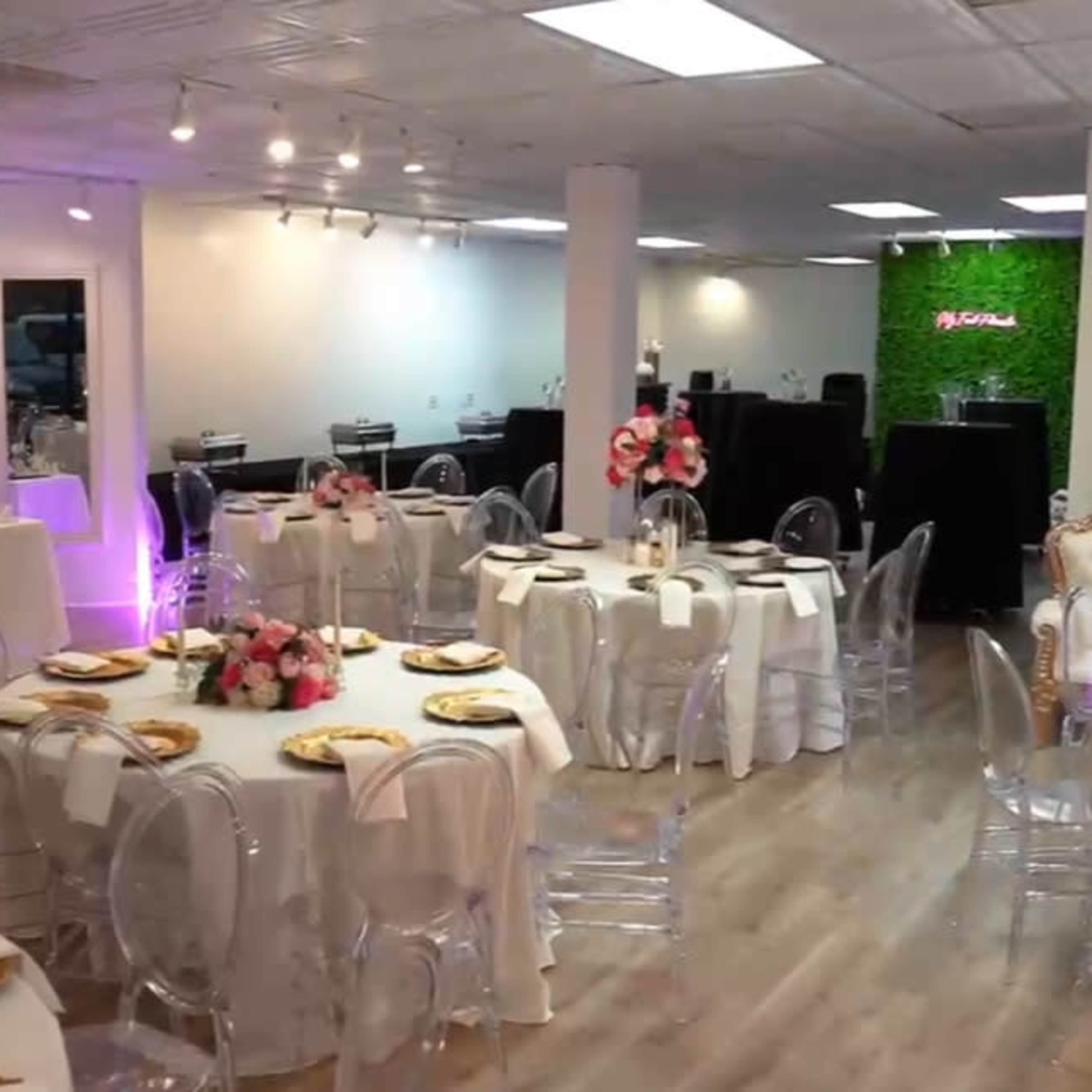 The room features elegantly set dining tables with clear chairs, floral centerpieces, and a backdrop adorned with decorations for a special event.