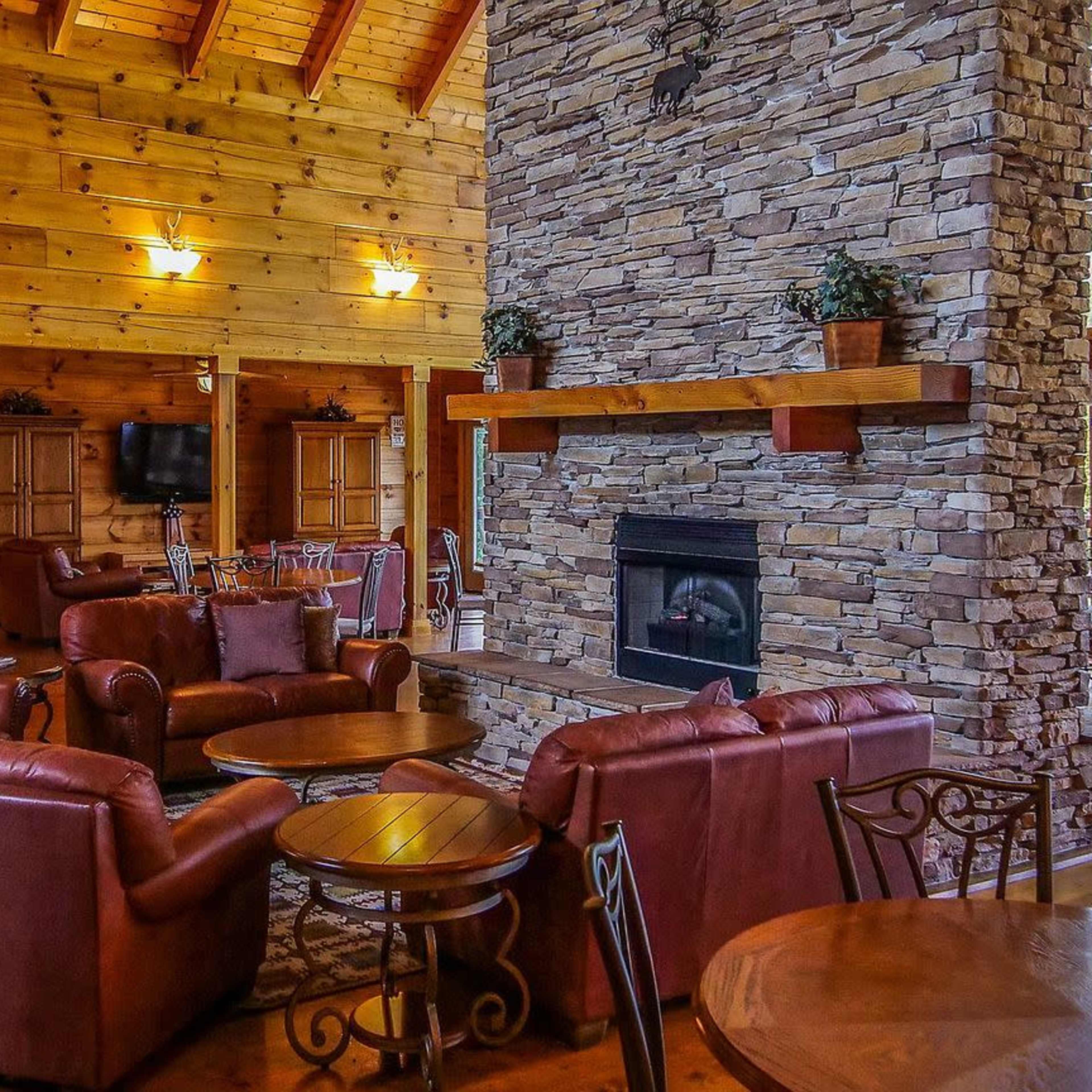 A spacious wooden interior of a cabin featuring a large stone fireplace, leather armchairs, and a dining area with wooden tables.