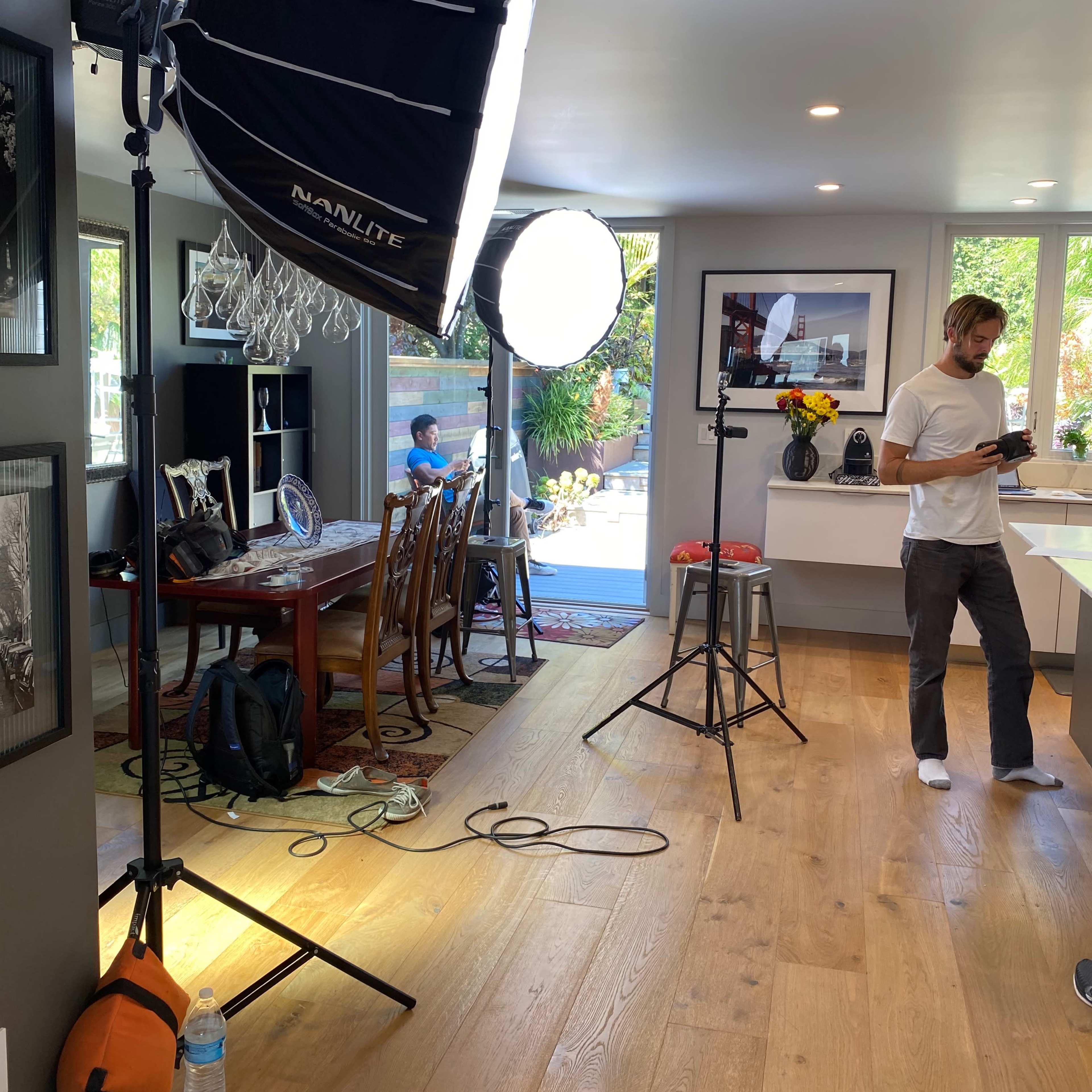 A well-lit interior scene shows photography equipment set up in a spacious dining area, with two people present and large windows providing natural light.