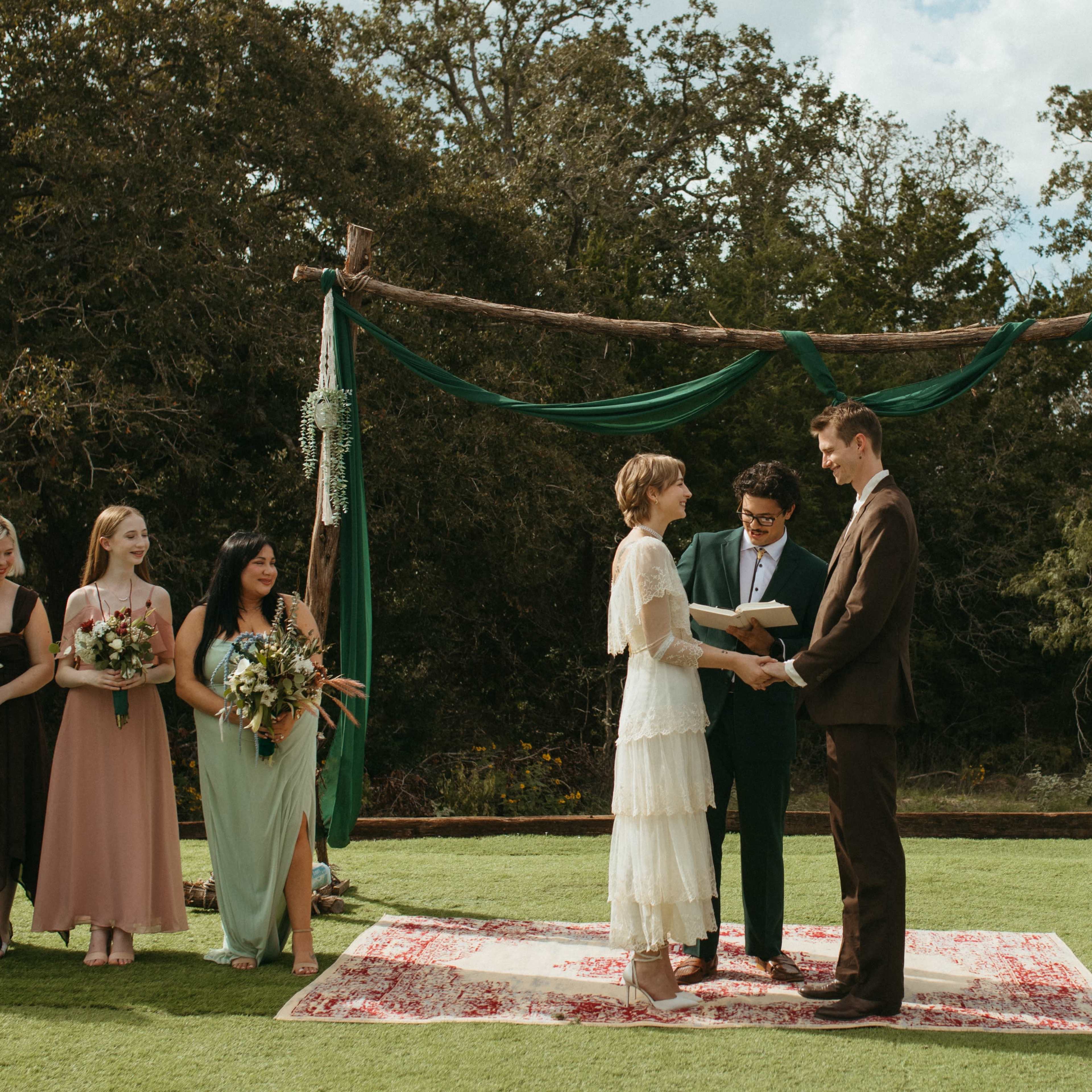 A couple stands under a wooden arch adorned with fabric, exchanging vows in front of their bridal party on a grassy outdoor setting.