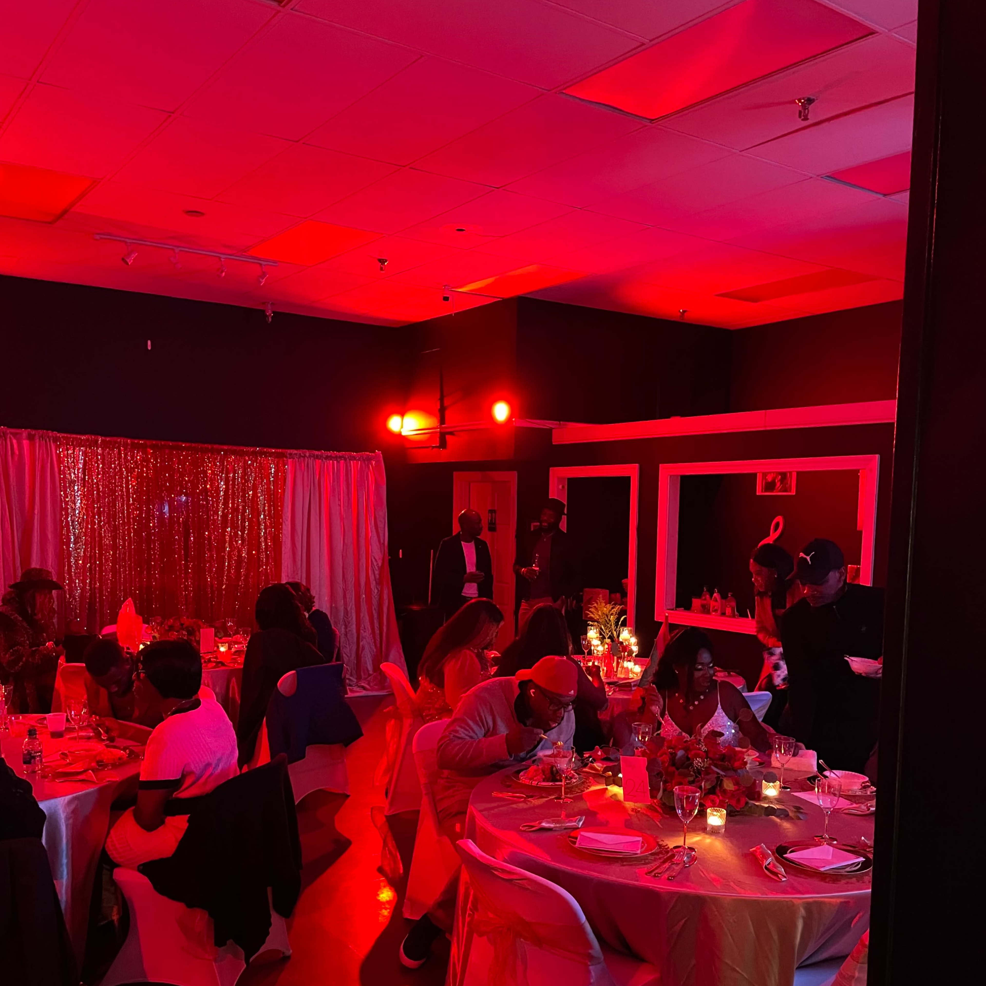A dimly lit event space with guests seated at elegantly set tables, illuminated by red lighting and decorated with shimmering backdrops.