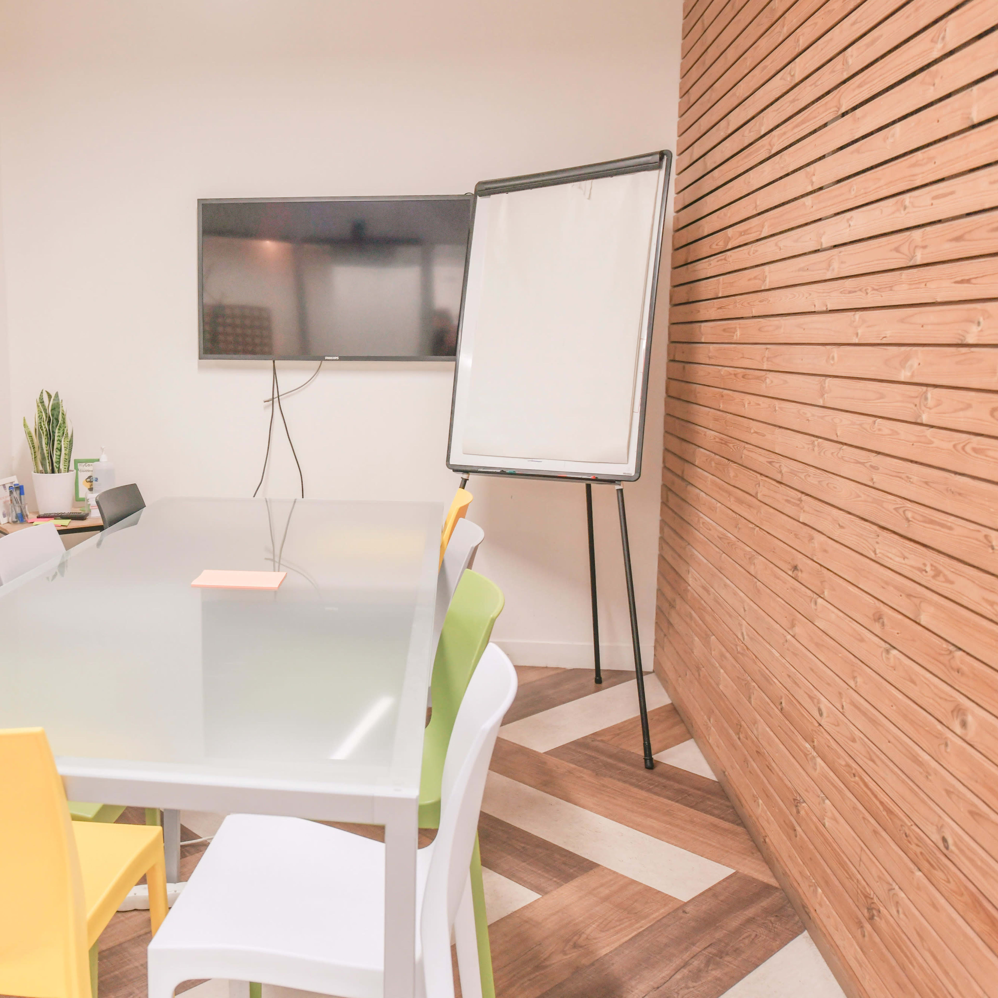 A conference room is set up with a glass table, colorful chairs, a wall-mounted television, and a flip chart on a stand.