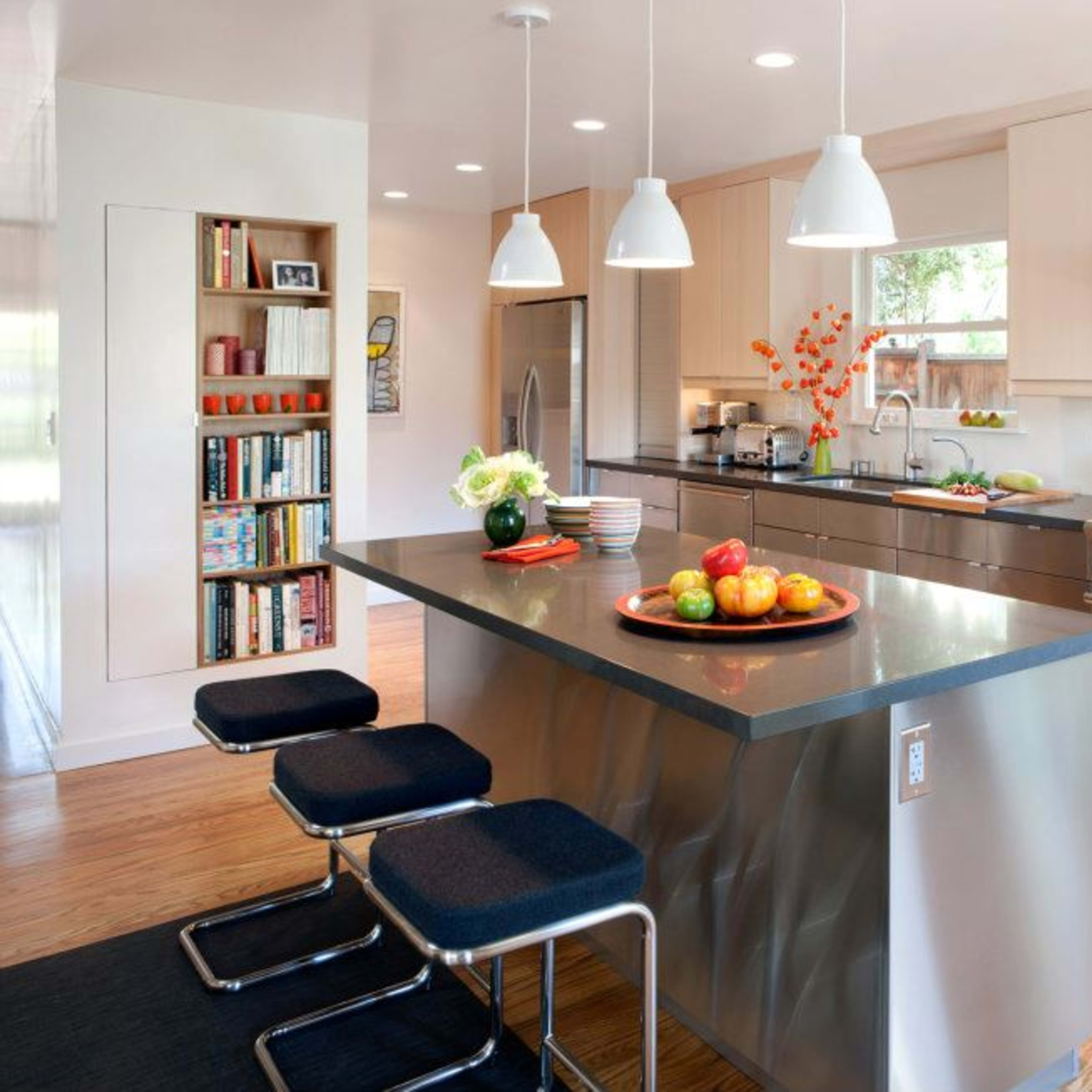 A modern kitchen features stainless steel appliances, a central island with a bowl of apples, and sleek cabinetry with a bookshelf along one wall.