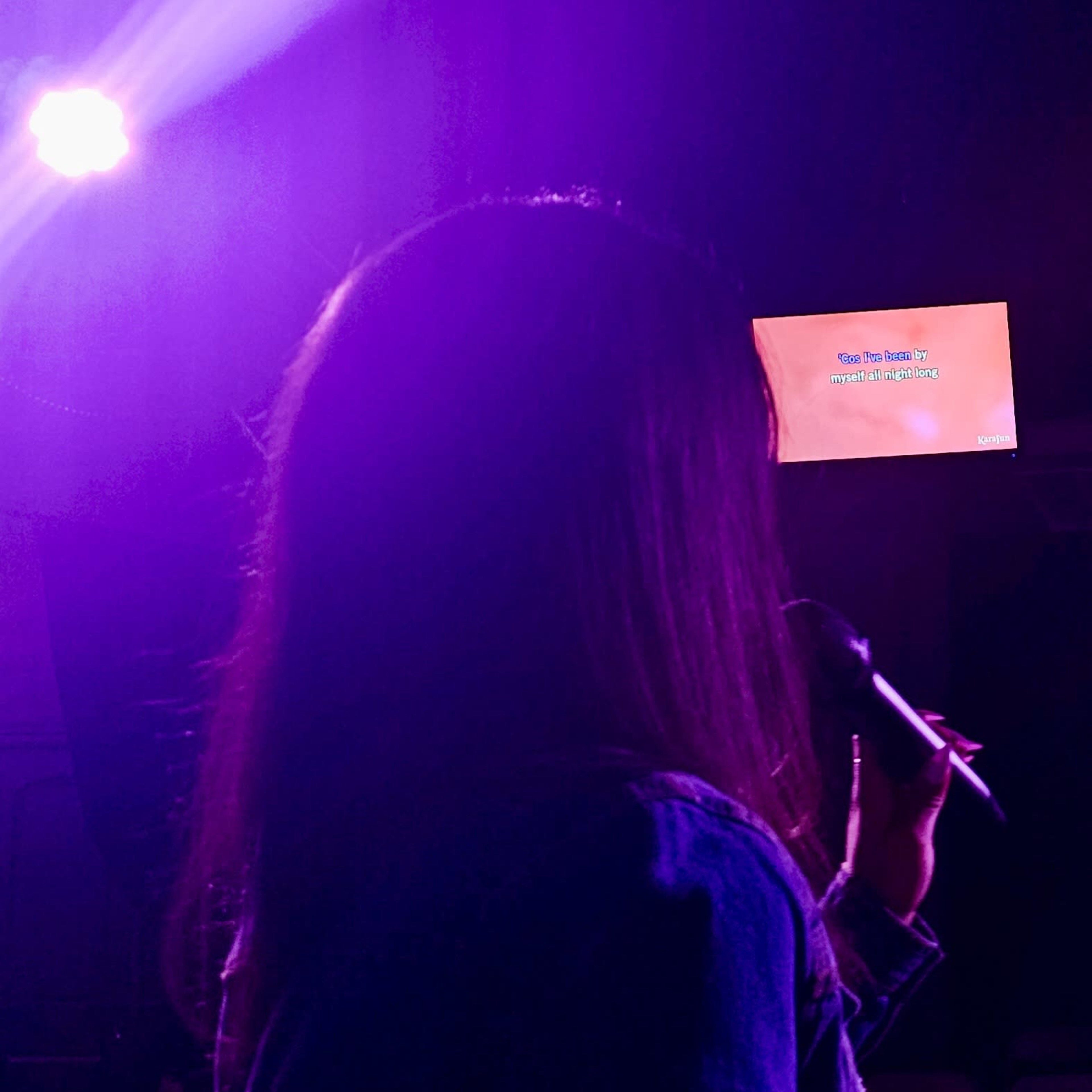 A person with long hair stands on stage holding a microphone, with a screen displaying lyrics in the background.