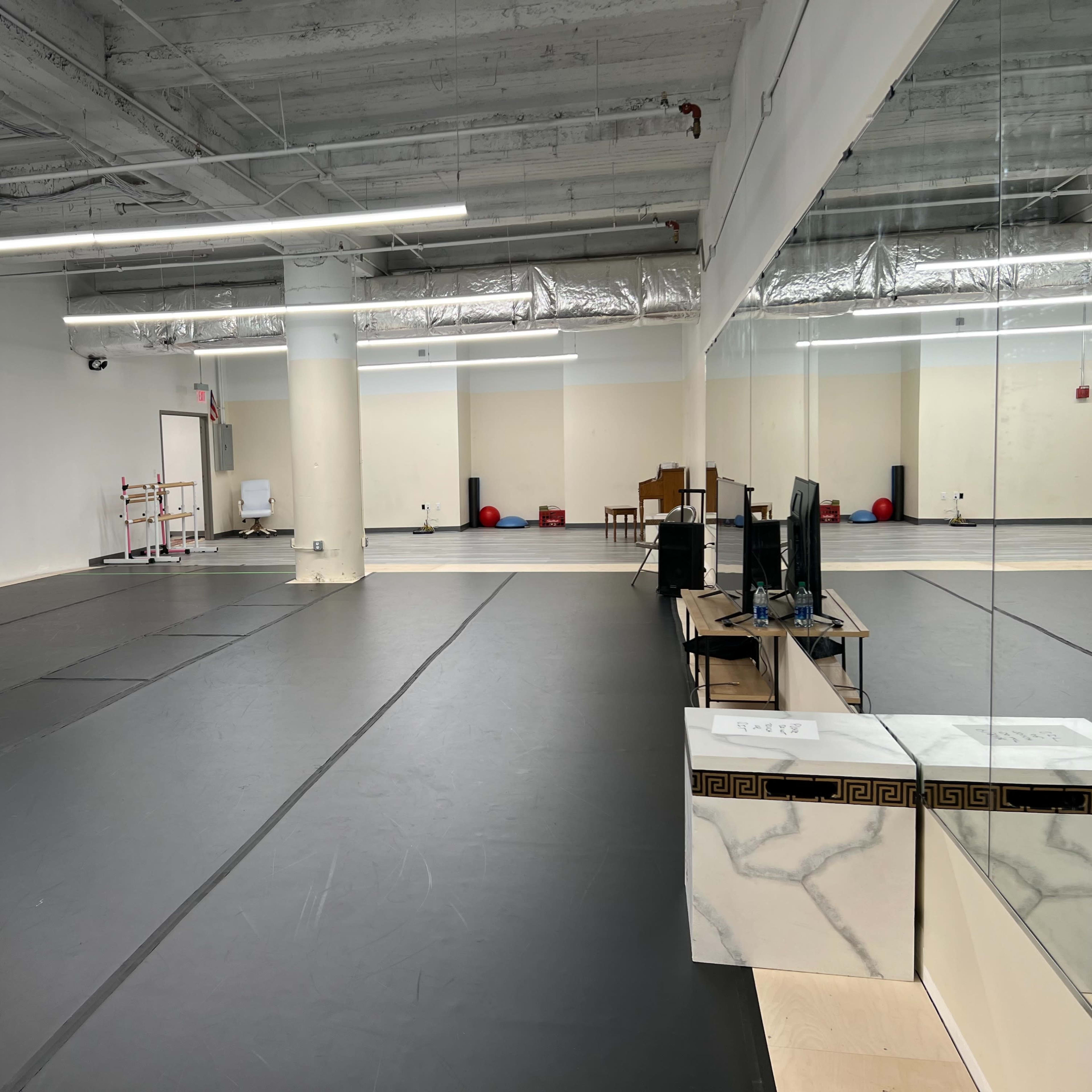 An empty gymnasium or studio with black mats, large columns, and mirrors along one wall.