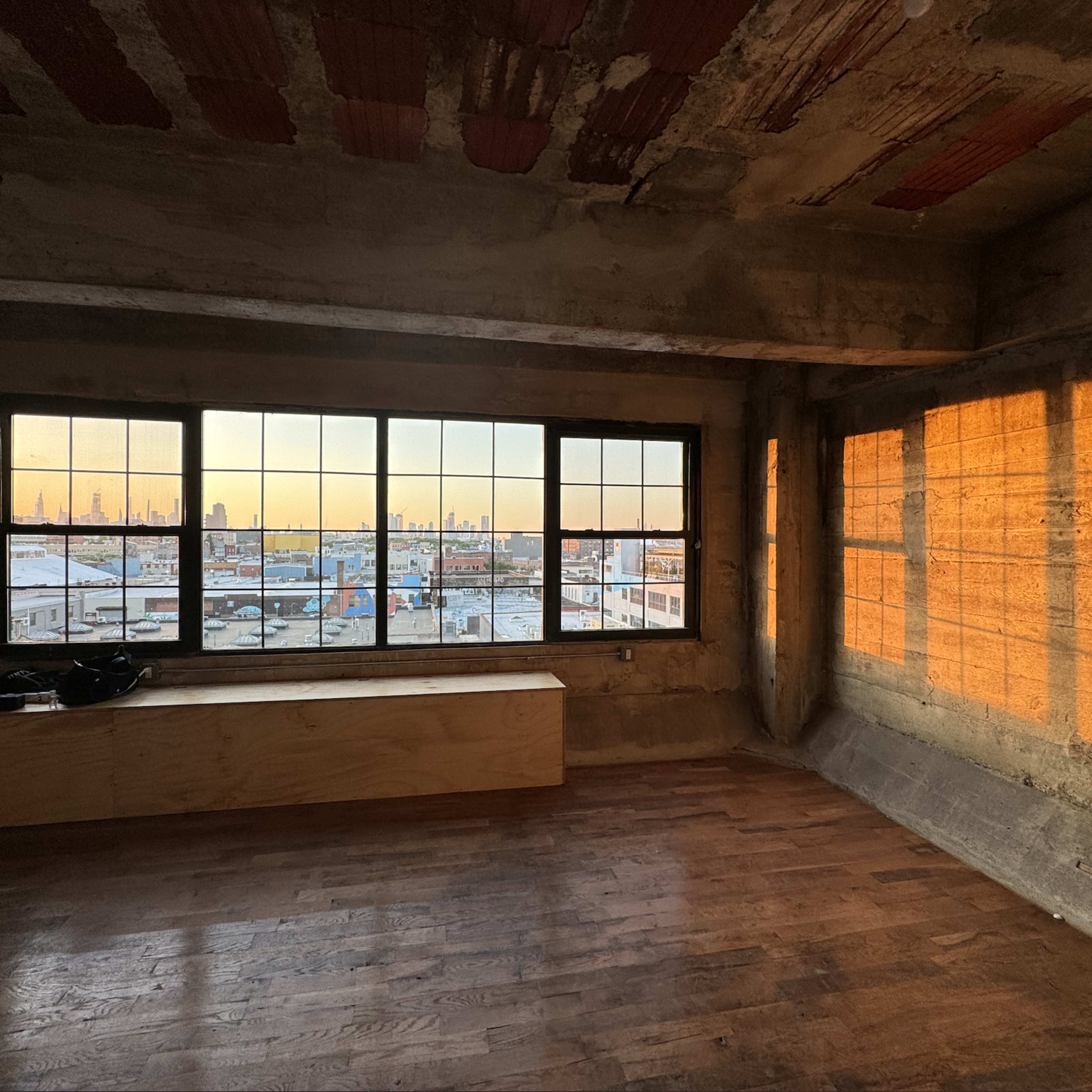 An unfinished interior room with large windows, allowing a view of a city skyline at sunset.