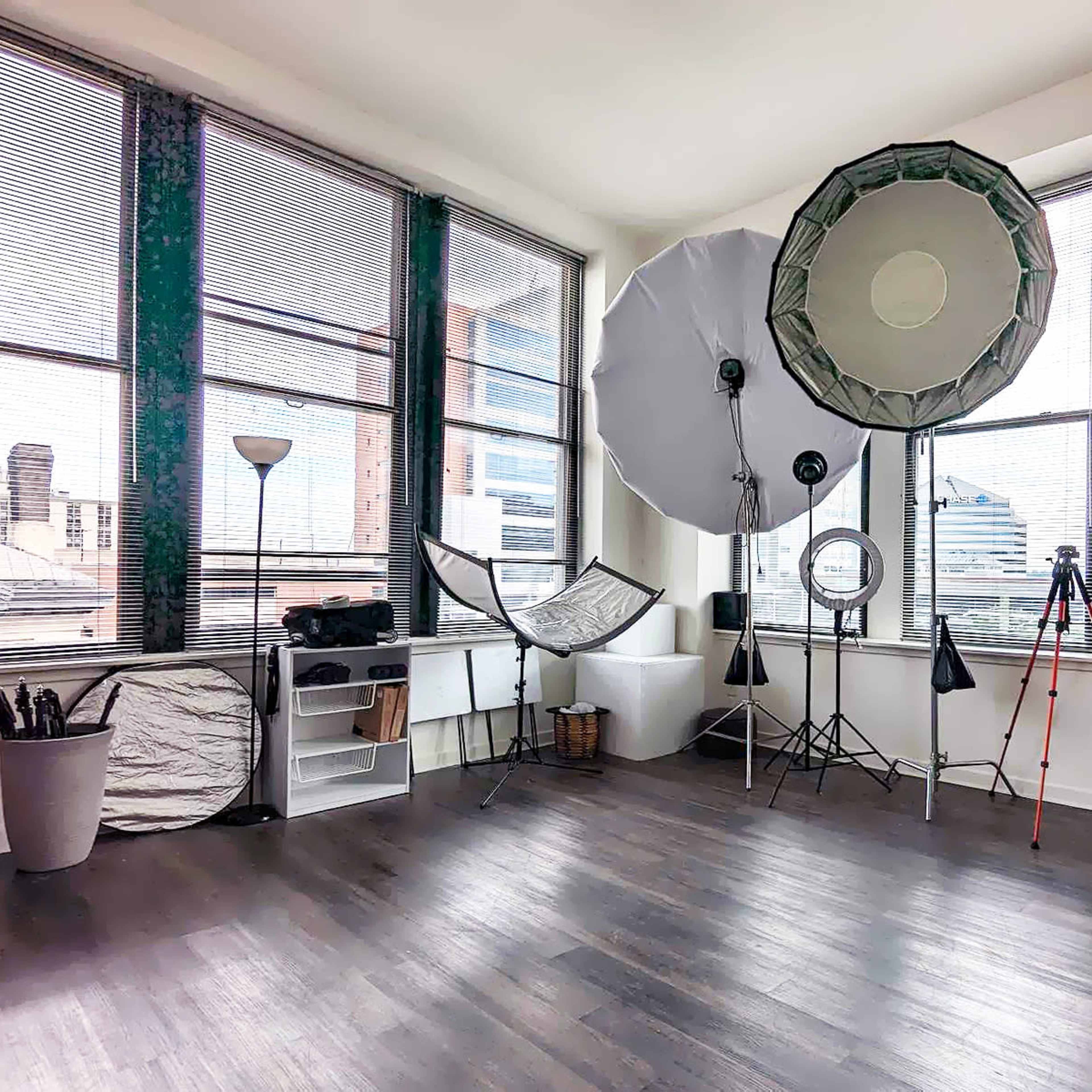 A spacious room with large windows, photography equipment including softboxes and tripods, and various items hanging on hooks.