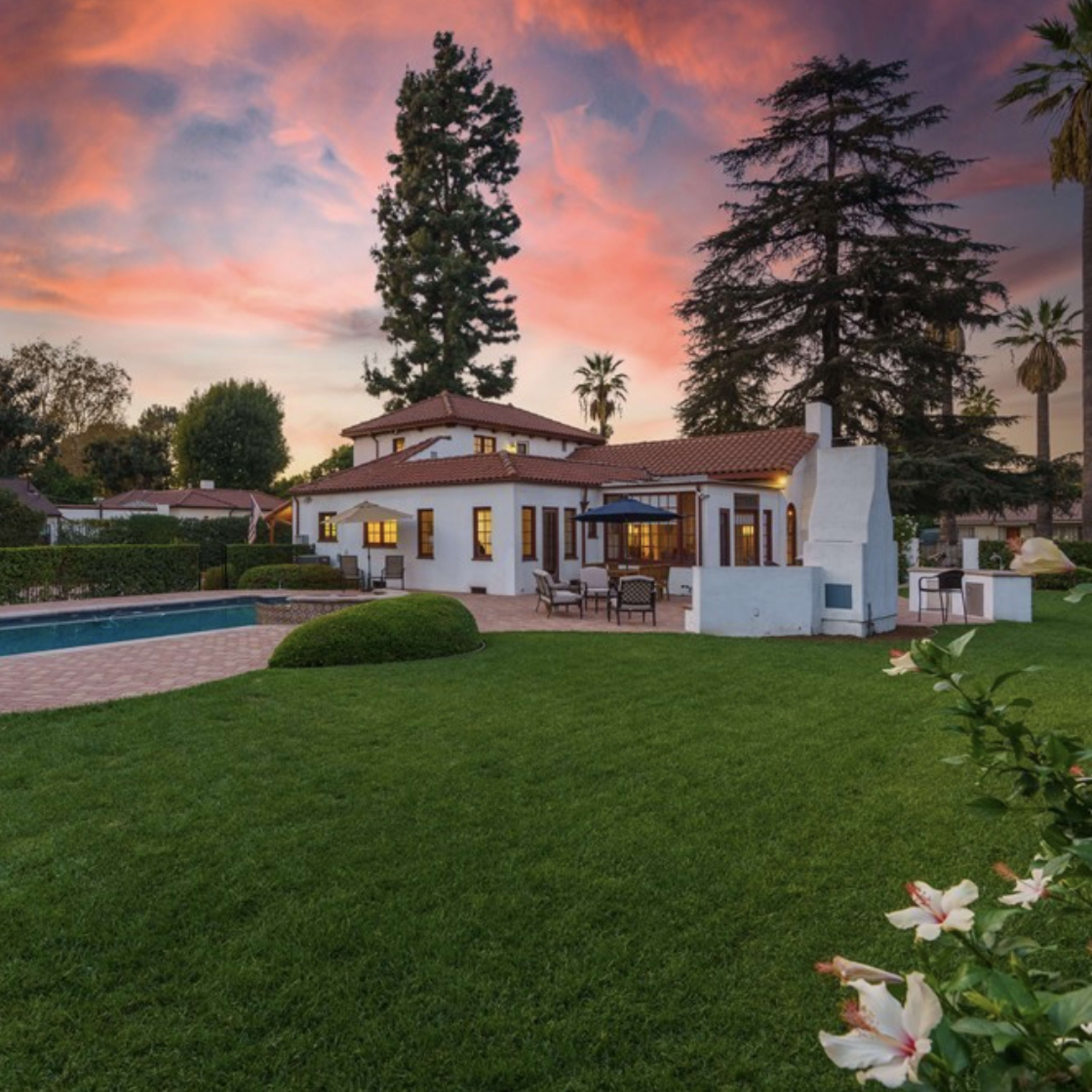 A backyard featuring a swimming pool, lush green grass, and a white house with red-tiled roofing, set against a vibrant sunset sky.