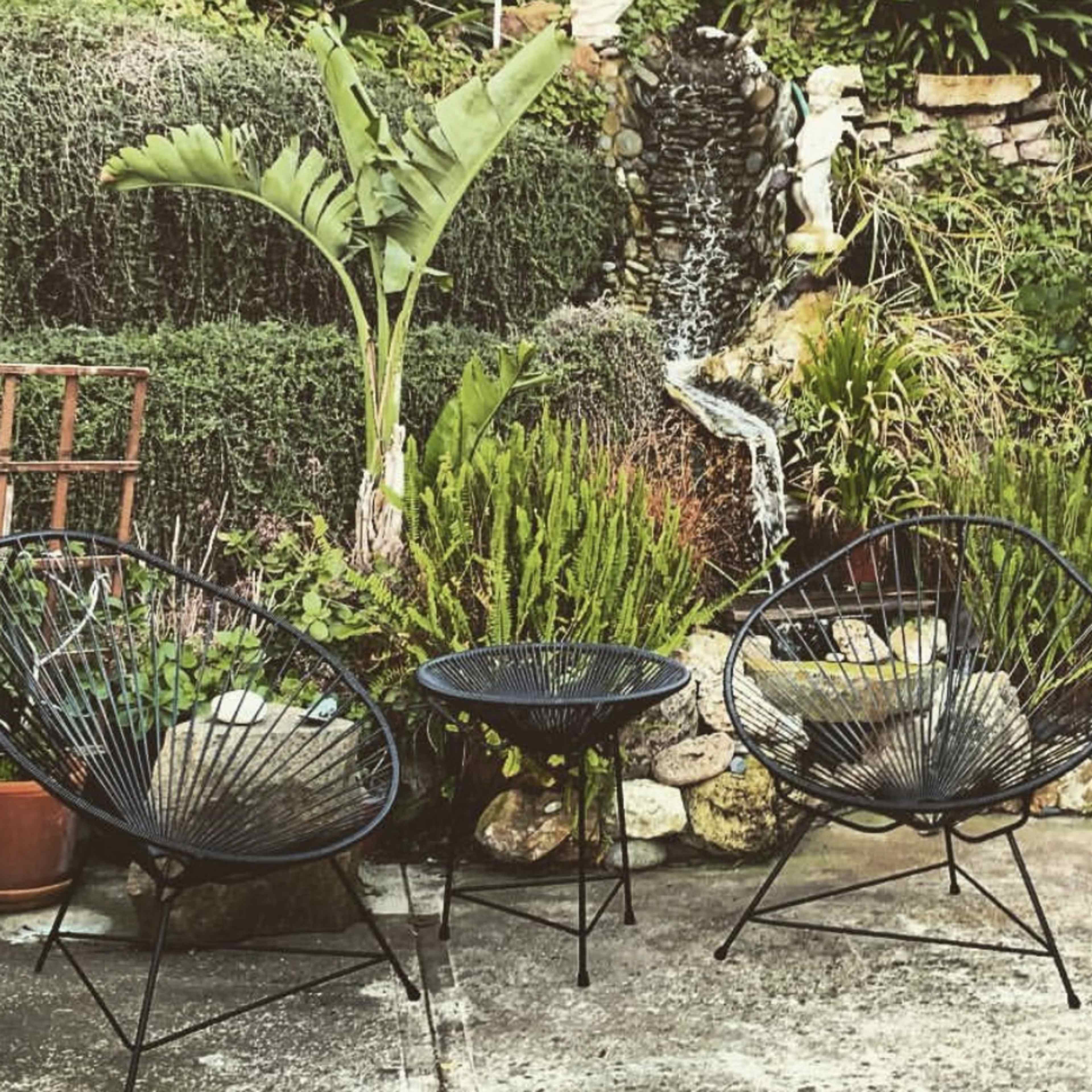 Two black wire chairs and a small round table arranged in a garden with a waterfall and lush greenery.