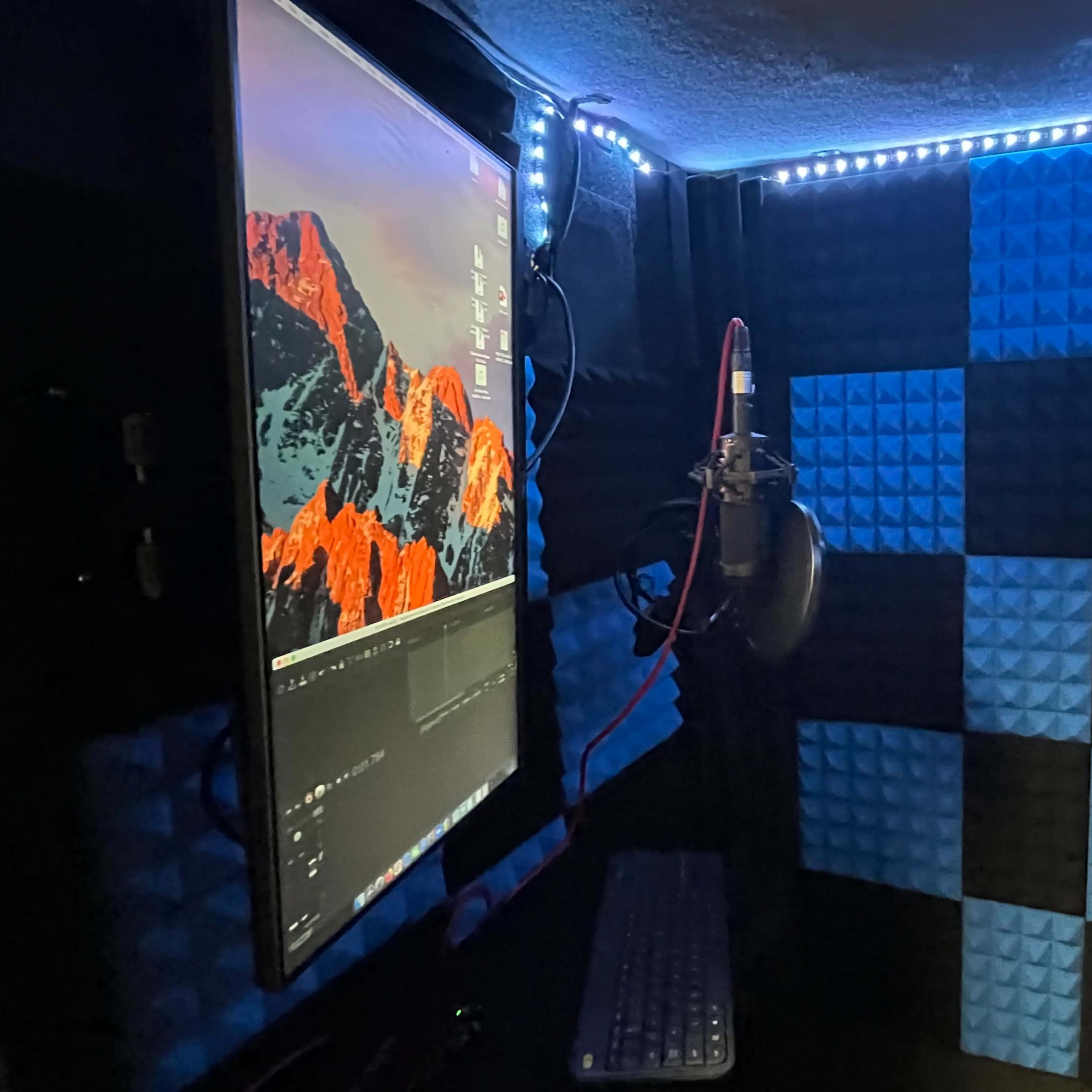 A recording studio booth featuring soundproof foam walls, a computer monitor, a keyboard, and a microphone.