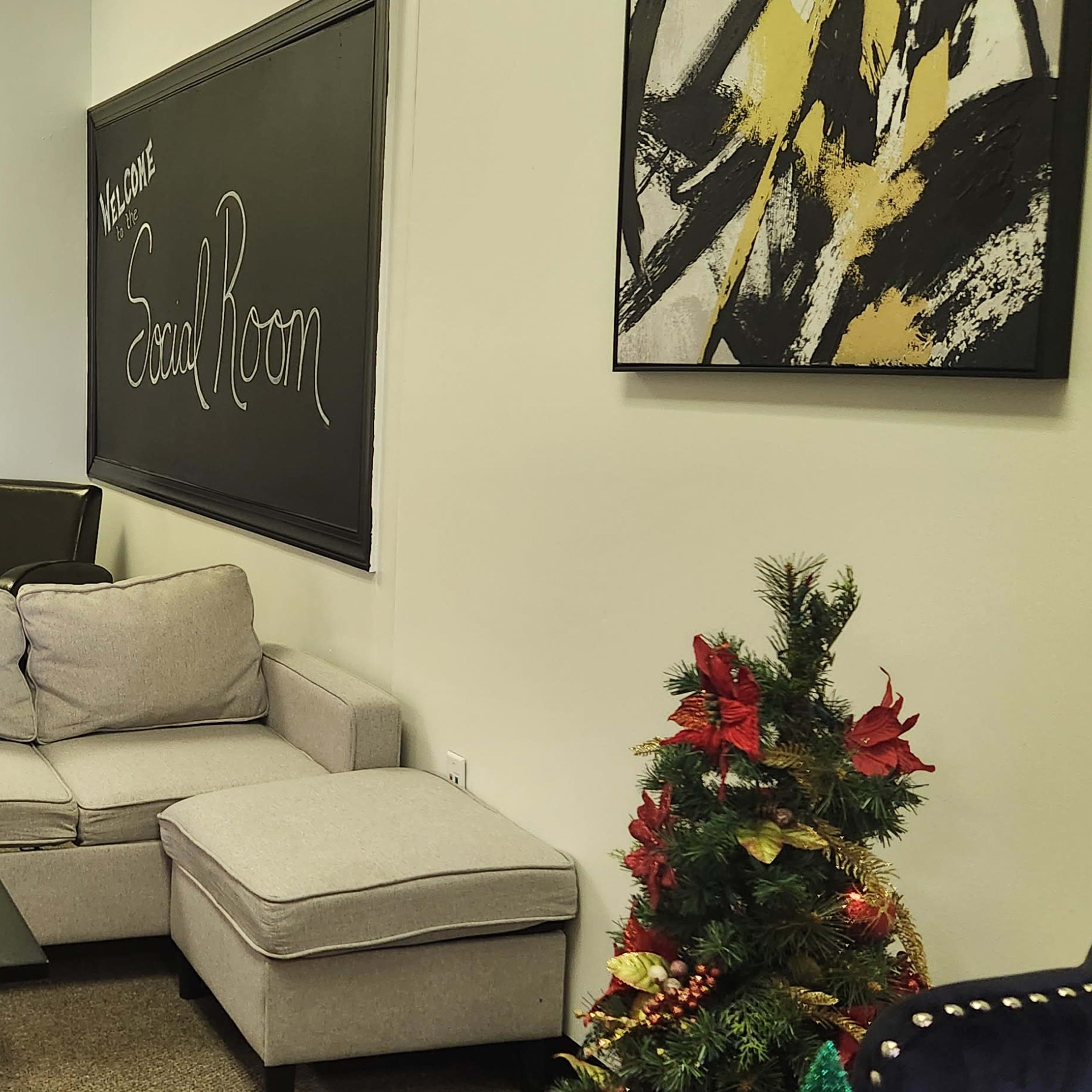 A waiting room with a gray couch, a small black table, and two black armchairs, decorated with artwork and a small festive plant.