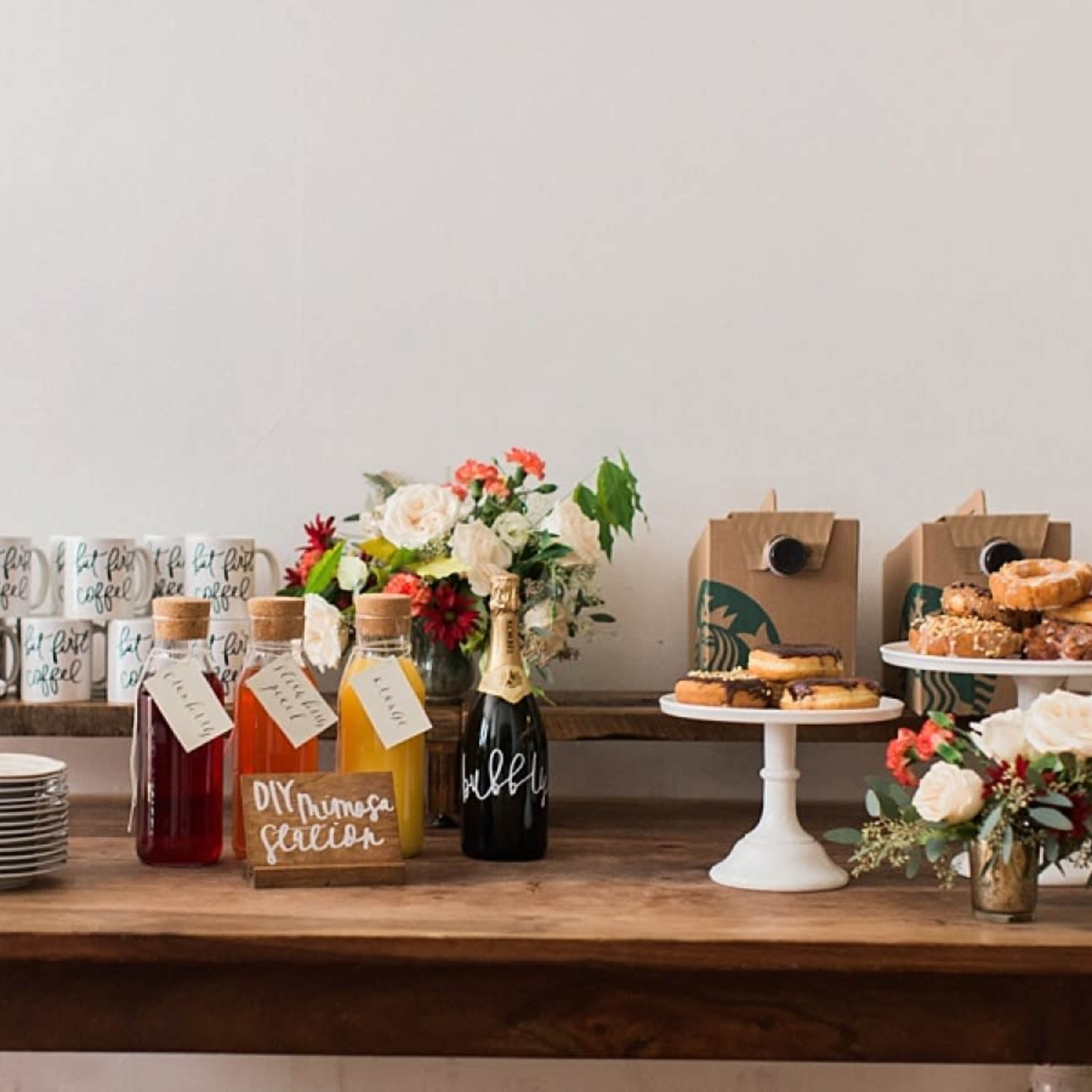 A wooden table is set with decorative bottles, baked goods, coffee mugs, and floral arrangements.