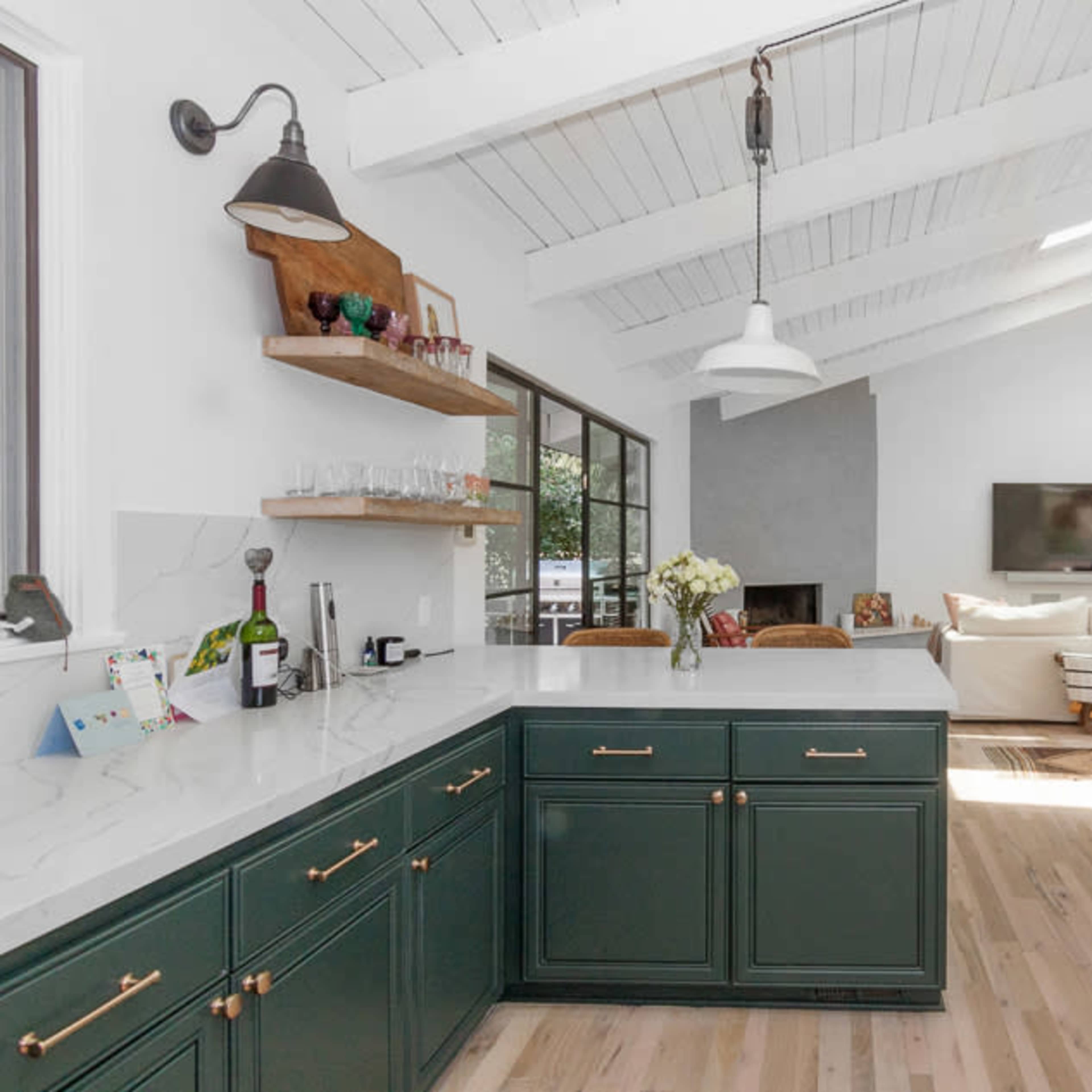 A modern kitchen features a green cabinetry with a white marble countertop, open shelving, and a bright living area in the background.