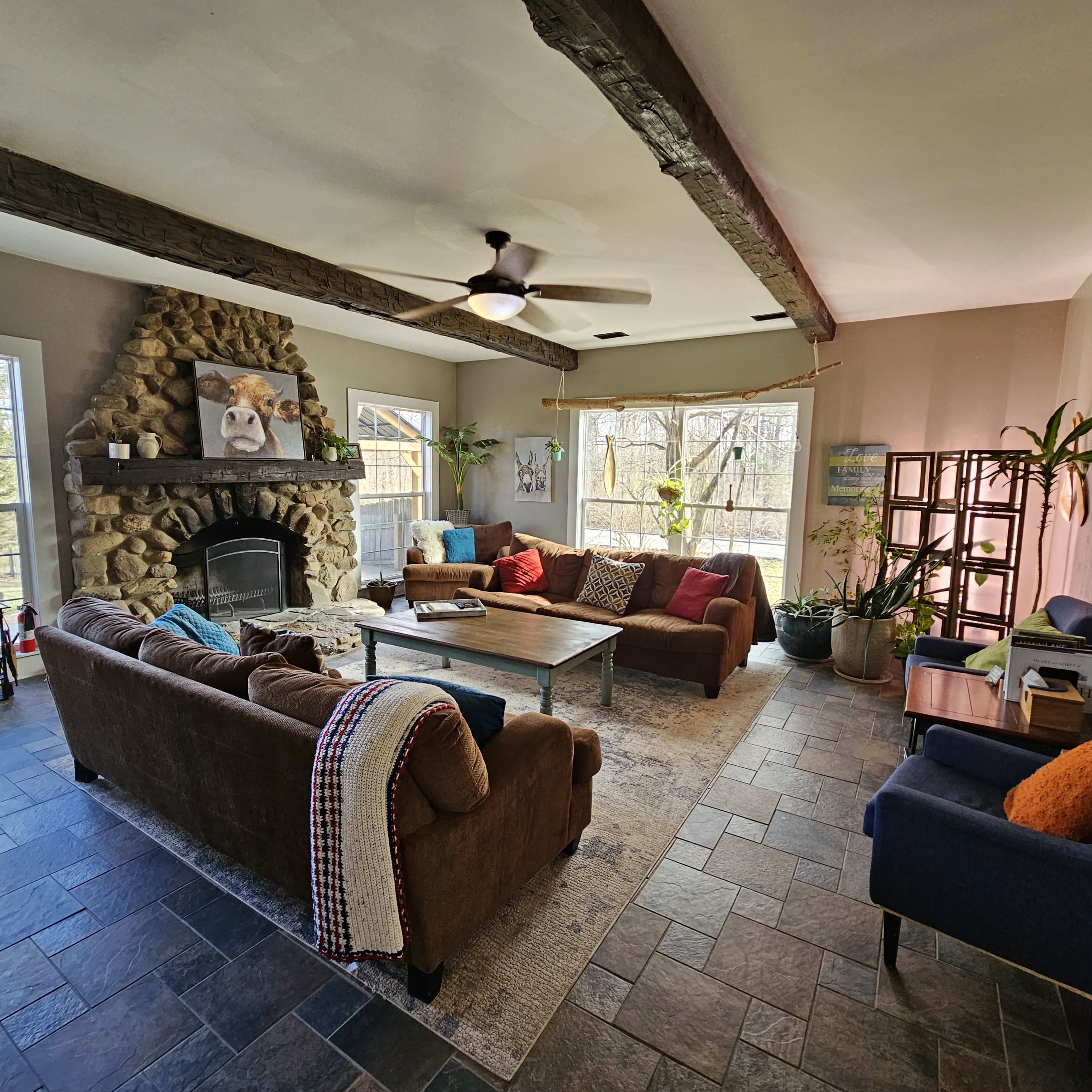 A spacious living room with a stone fireplace, two sofas, a coffee table, and large windows overlooking a wooded area.