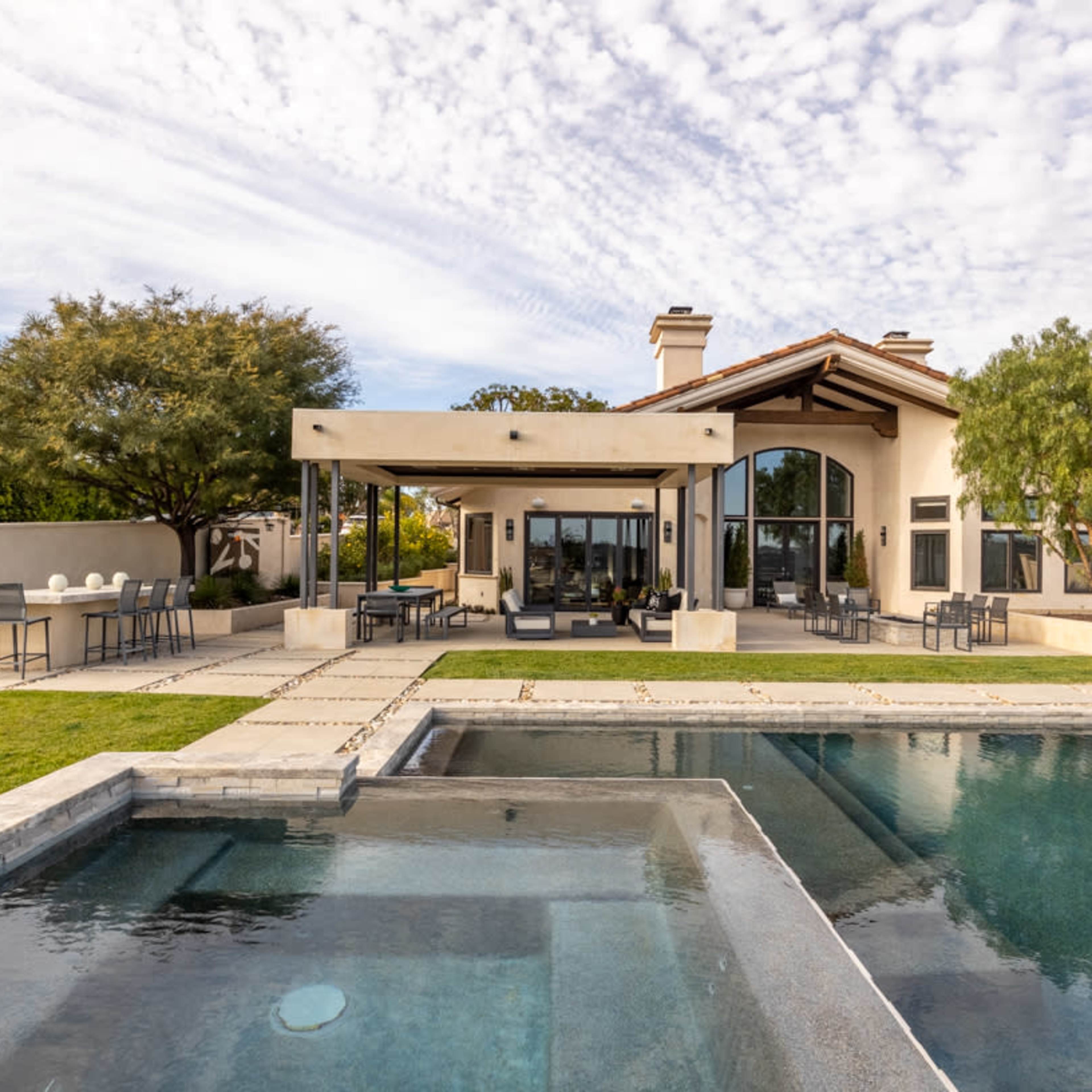 A modern house with a large patio, surrounded by landscaping and a swimming pool in the foreground.