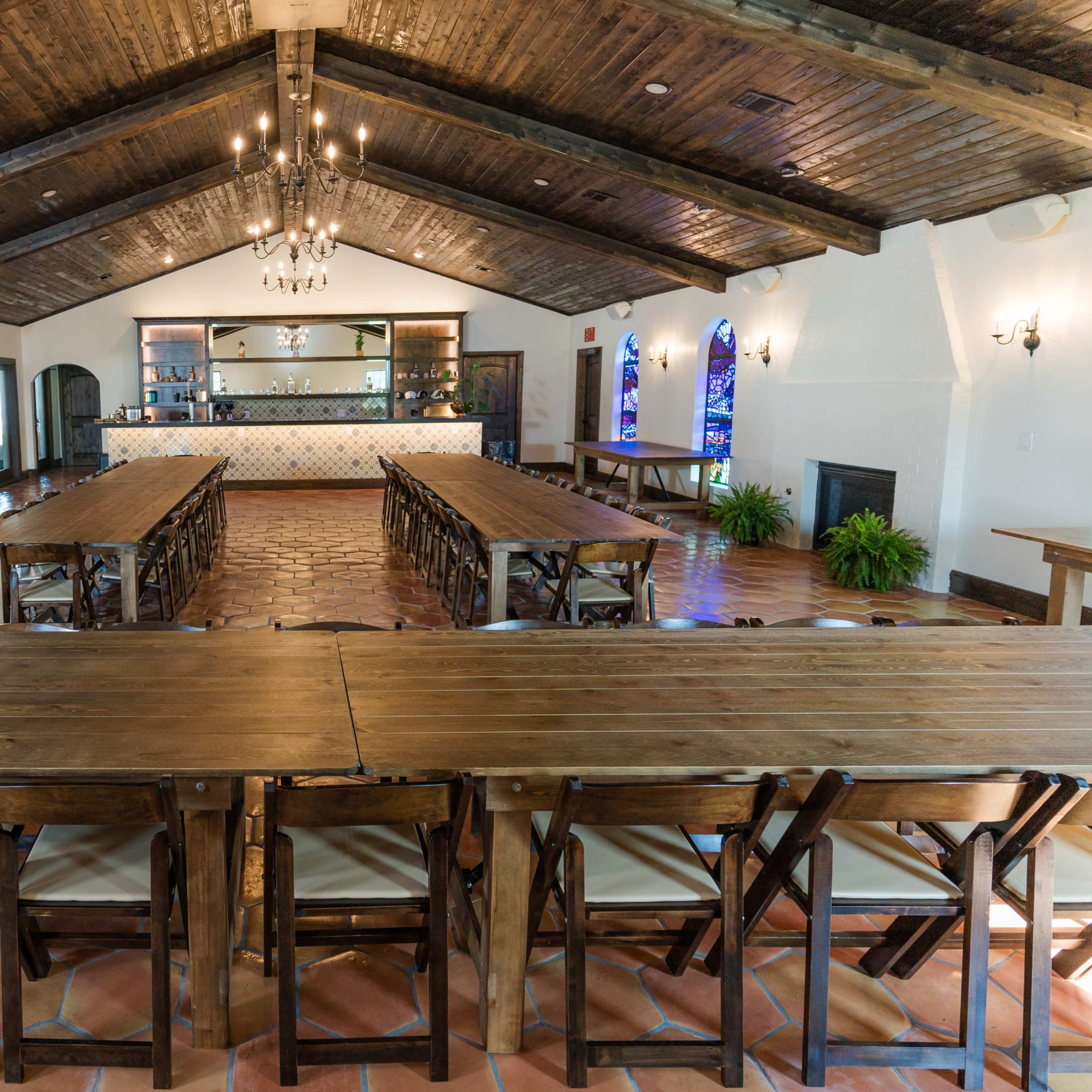 A spacious dining area with wooden tables arranged in a long configuration, featuring a bar with shelves at the back and pendant lighting above.