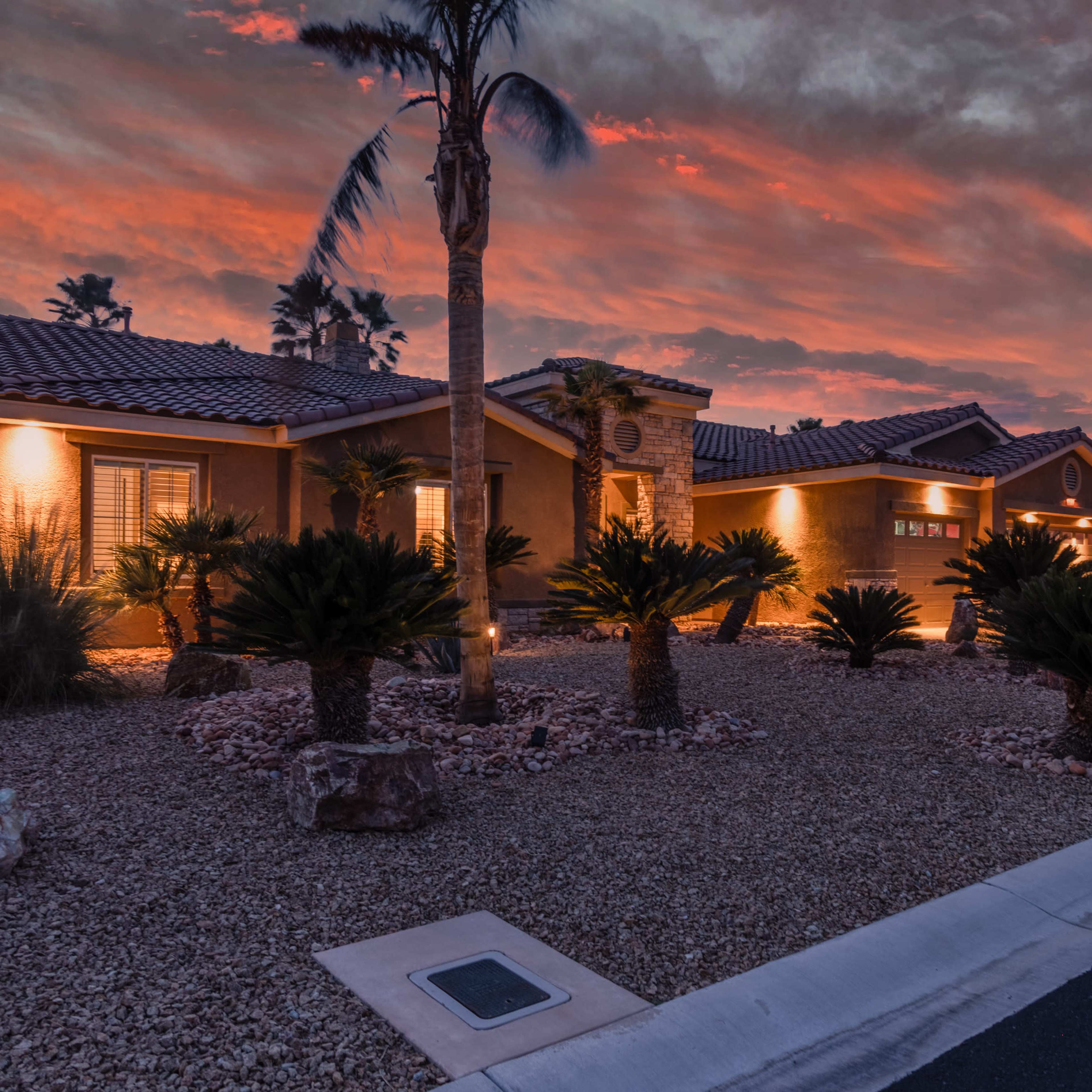 A row of houses illuminated by exterior lights, set against a colorful sunset sky with palm trees and landscaping featuring desert plants.