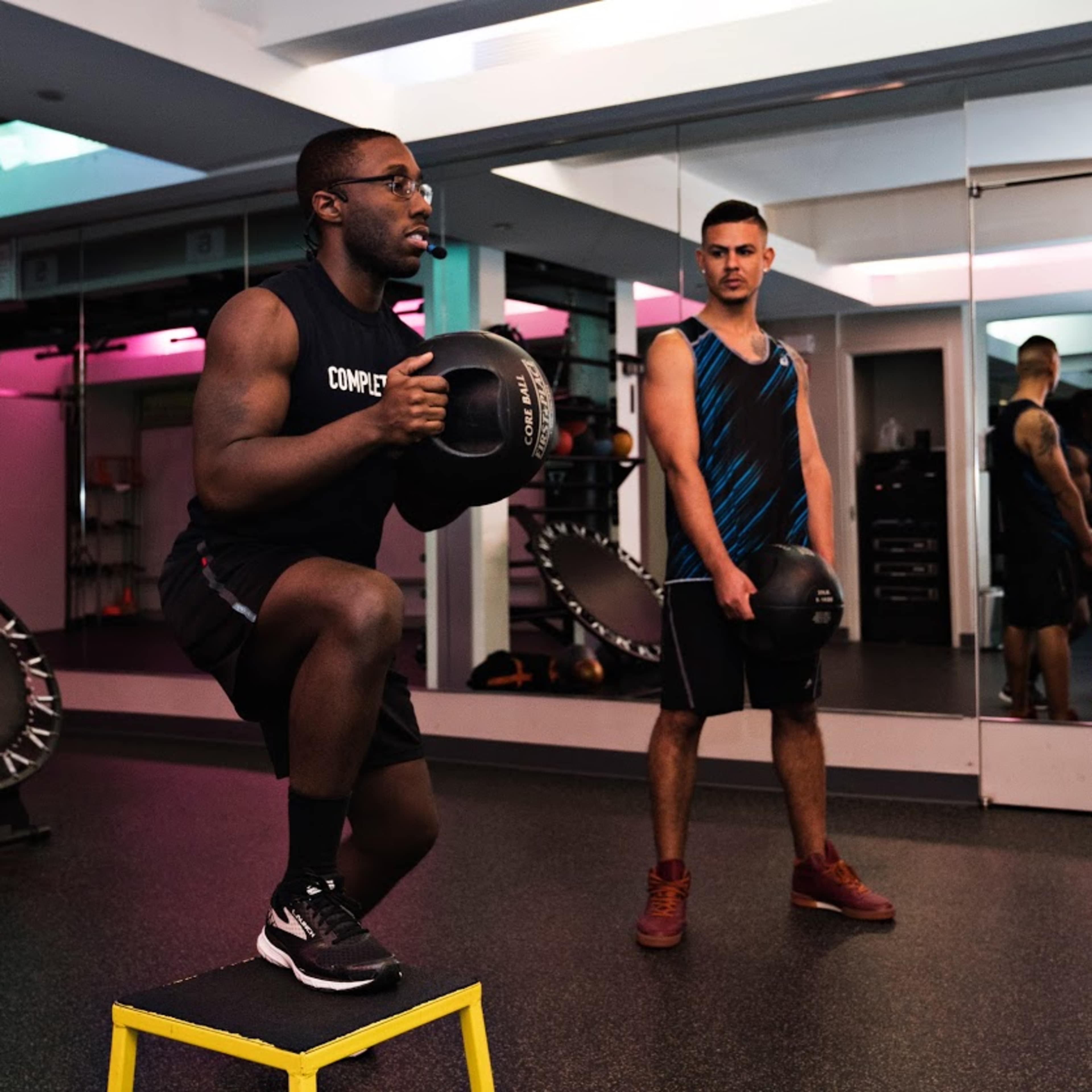 Two men are exercising in a gym, one performing a squat on a platform while holding a dumbbell, and the other observing nearby.