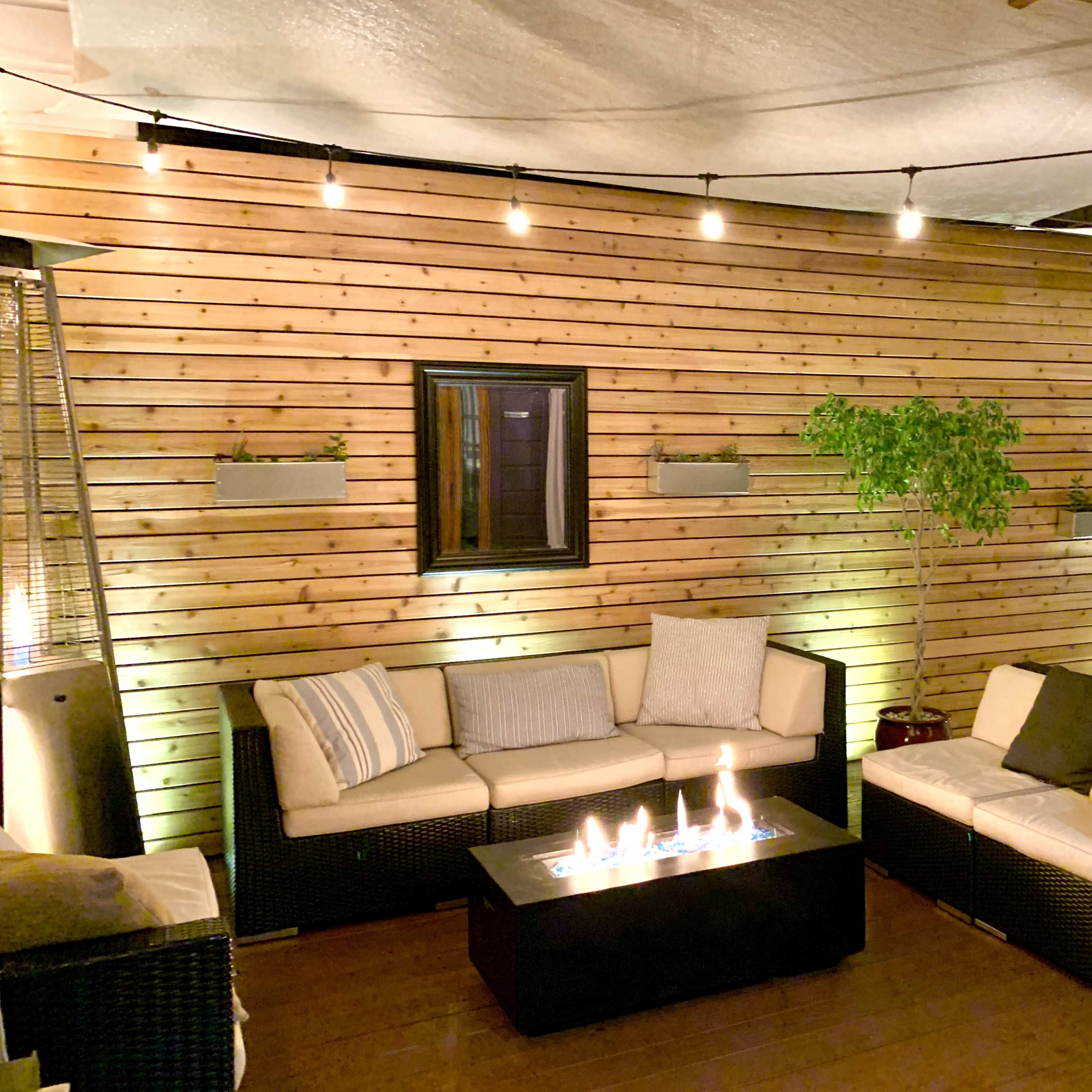 A cozy outdoor seating area with a wooden wall, a fire pit table, and string lights illuminating the space.