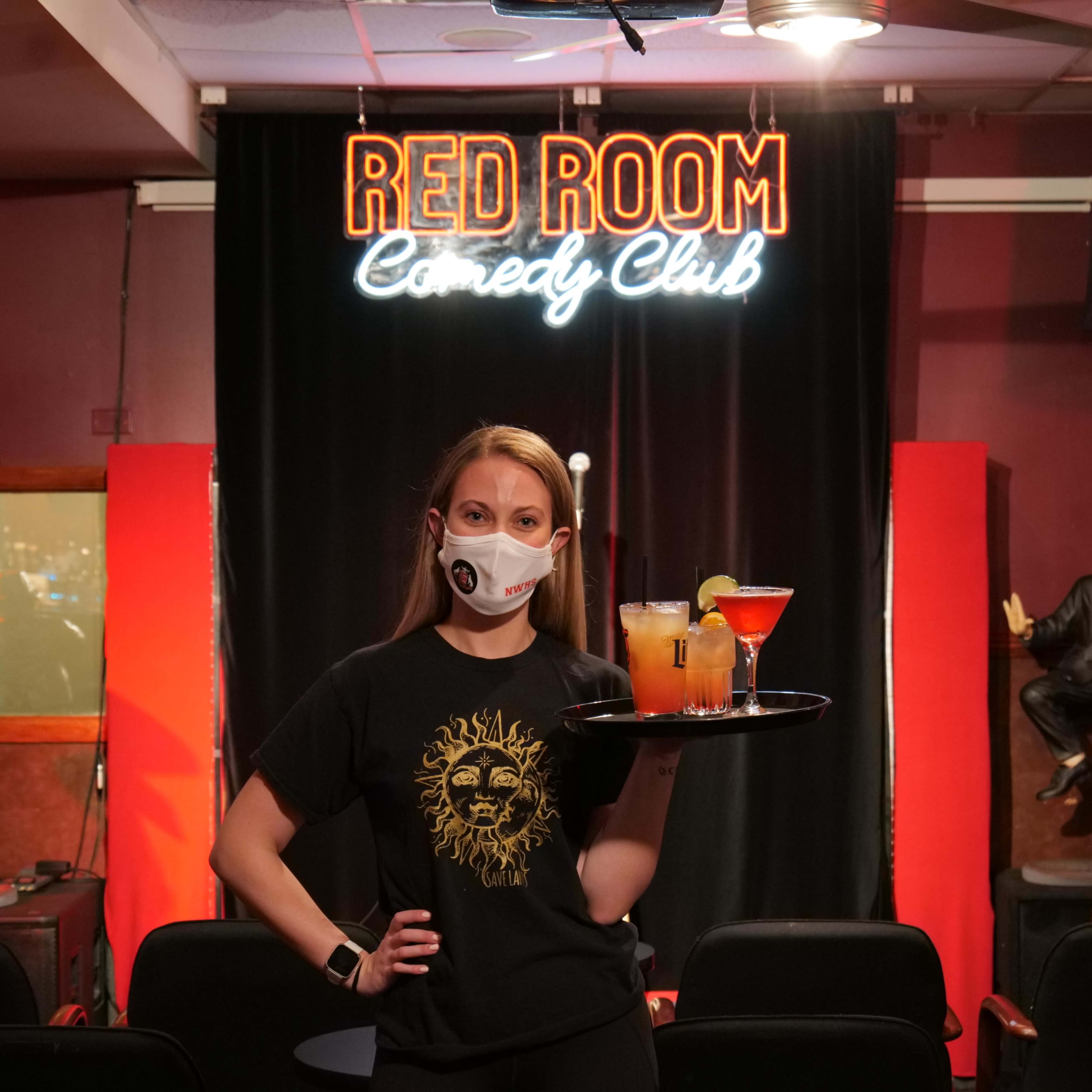 A masked waitress stands in front of the Red Room Comedy Club stage, holding a tray of drinks.