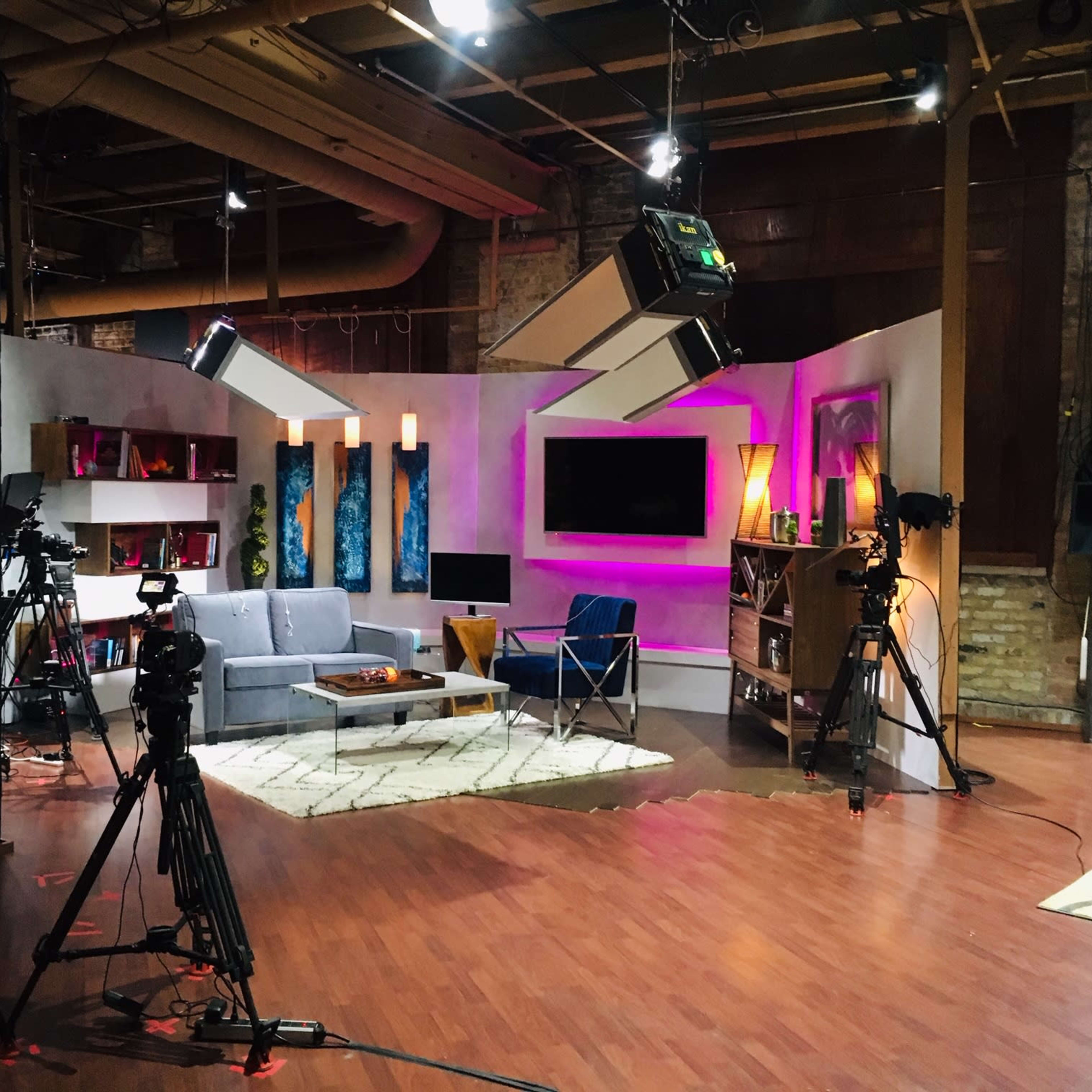 A television studio set with a gray sofa, colorful wall art, and multiple cameras positioned around a brightly lit area.