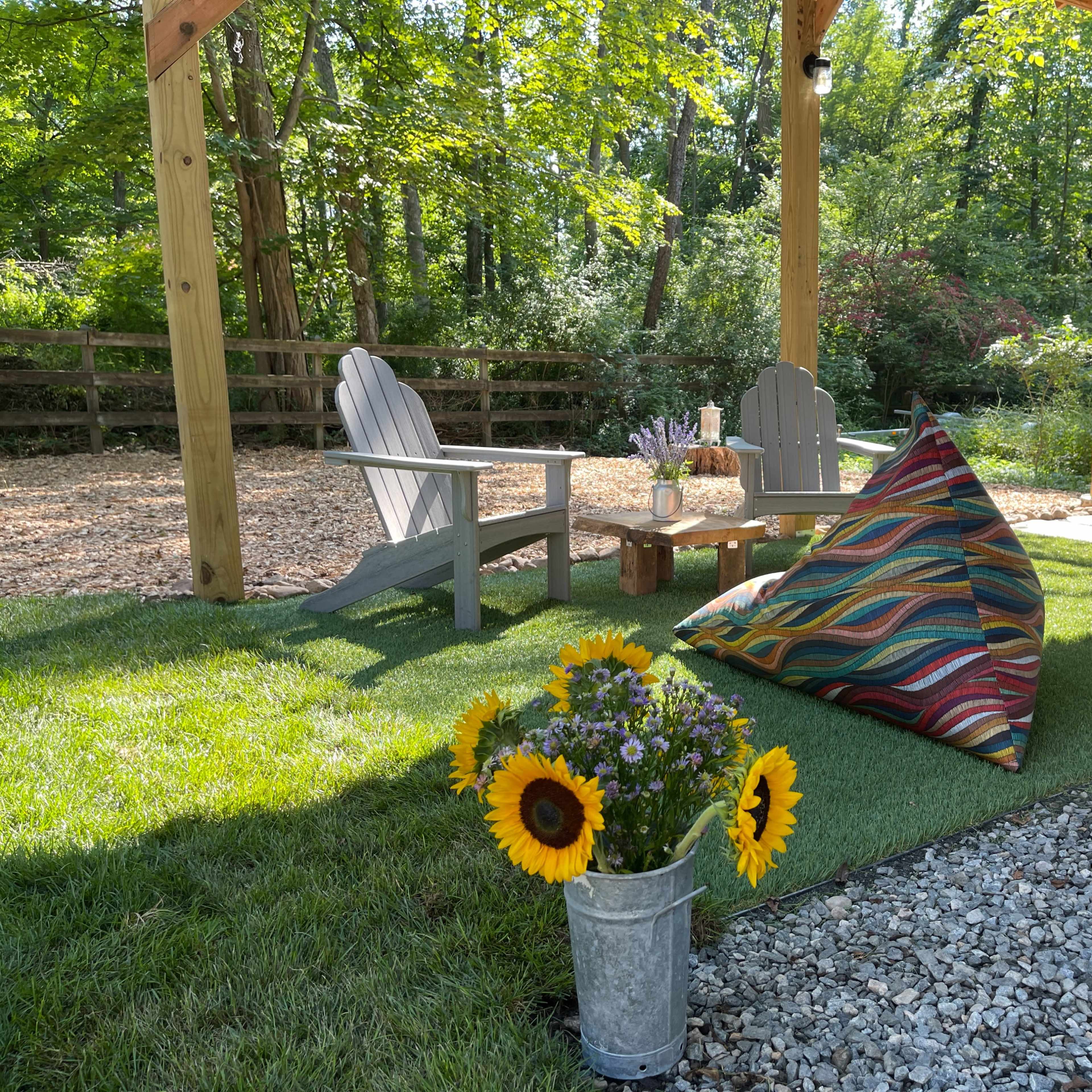 A cozy outdoor seating area features two Adirondack chairs, a colorful triangular cushion, and a bucket of sunflowers on a grassy mat under a wooden pergola.