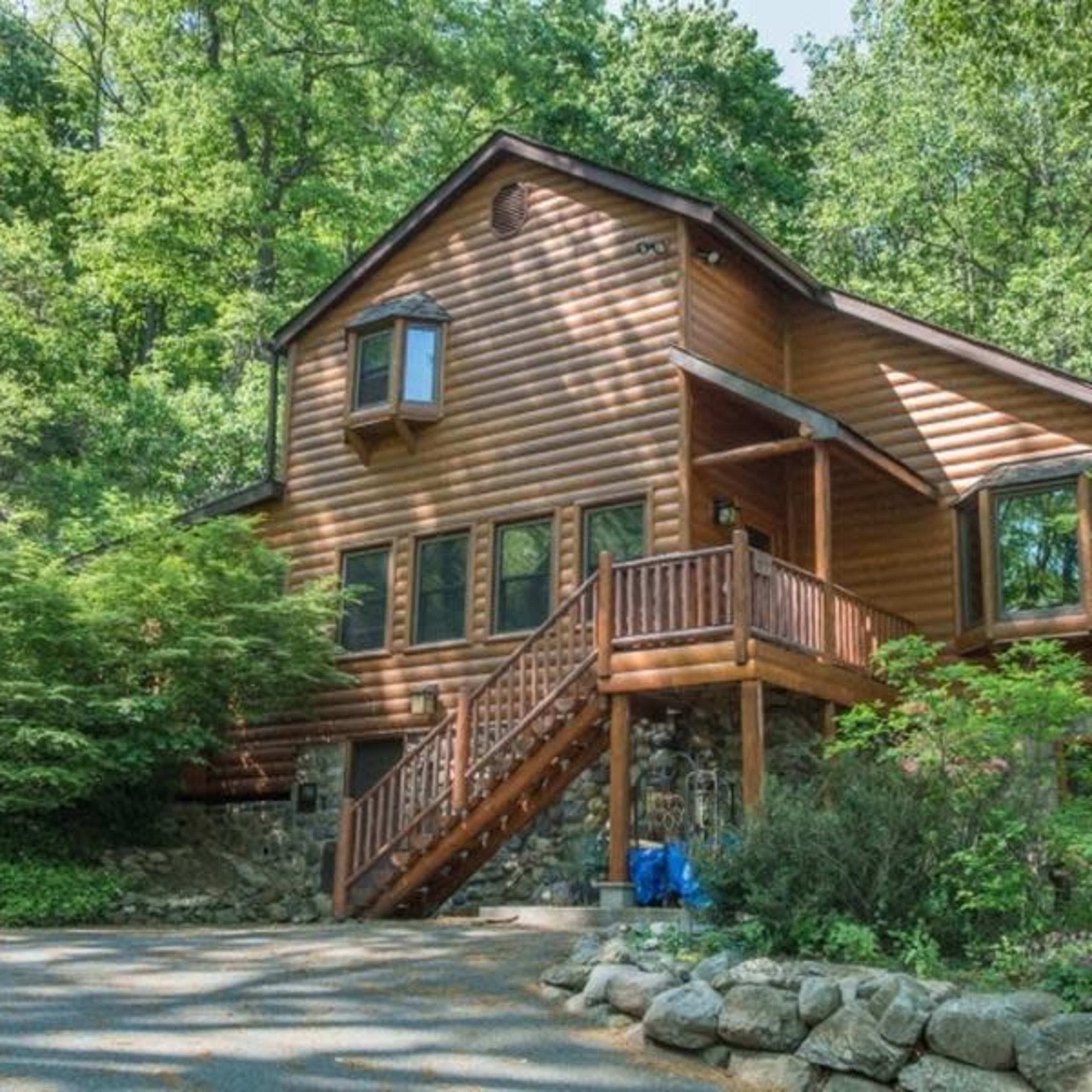A two-story wooden cabin nestled among lush green trees, featuring a front porch and stone accents.
