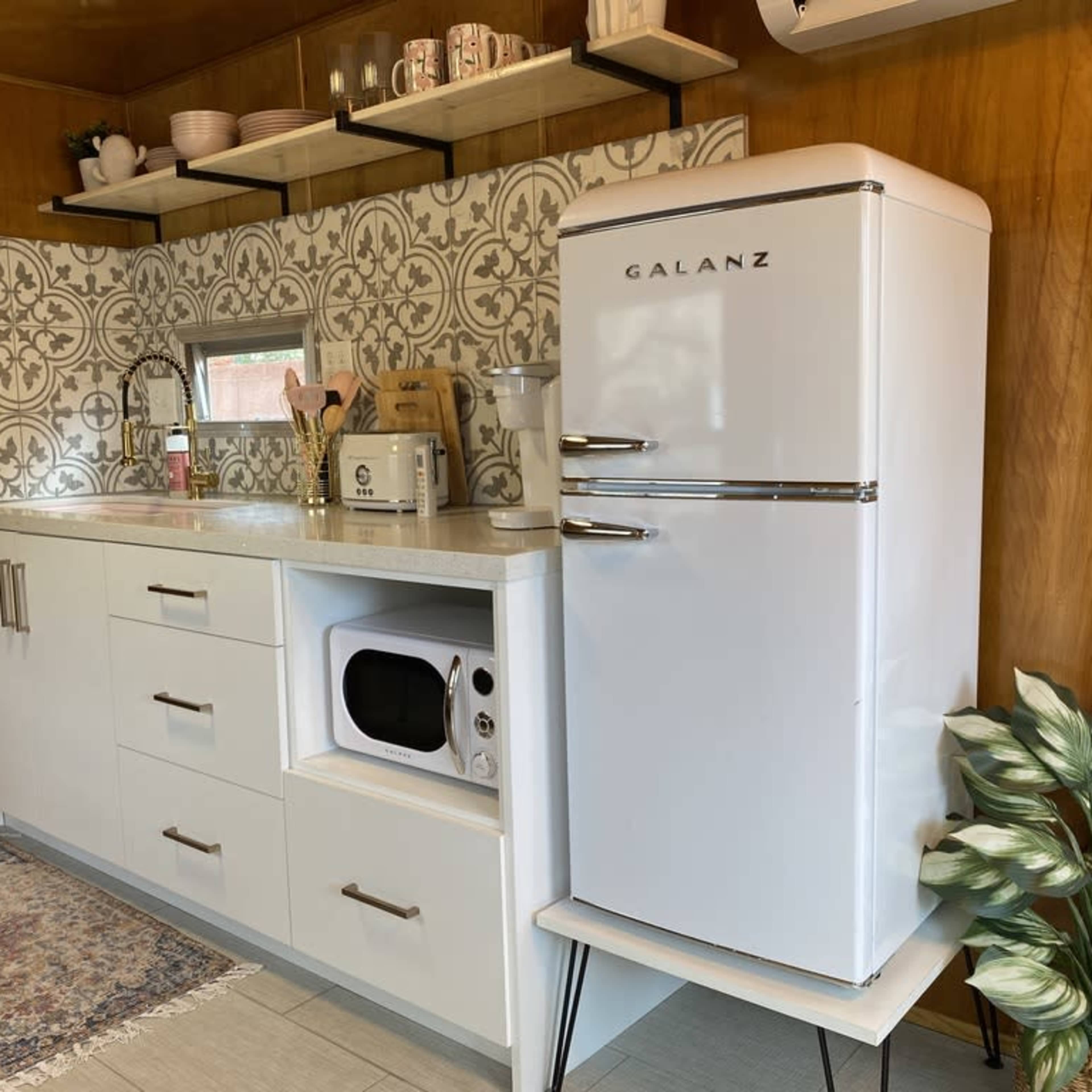 A small kitchen featuring a white retro-style refrigerator, a microwave under the counter, and patterned tile backsplash above a sink and countertop.