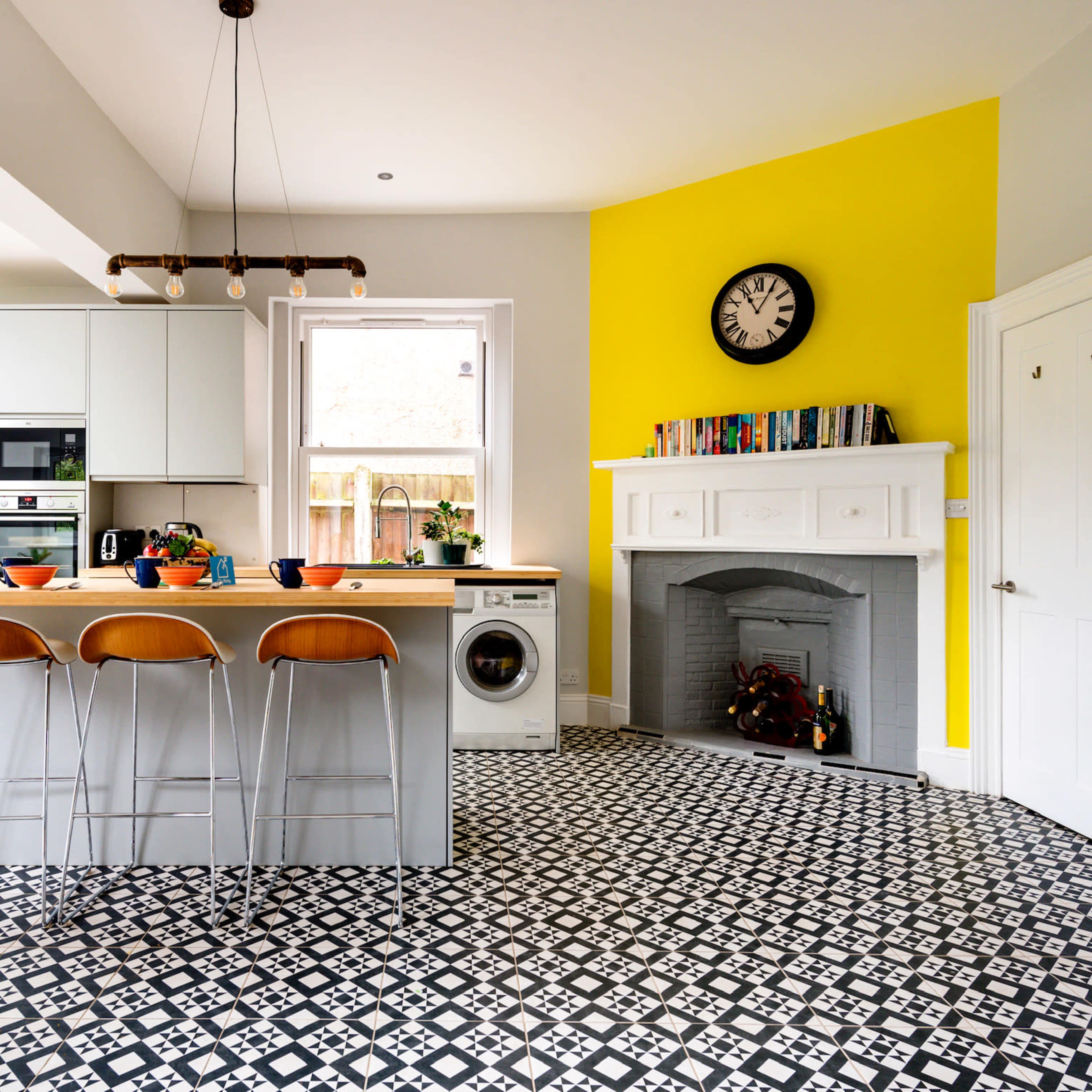 A modern kitchen with a yellow accent wall, a wooden island with three stools, and a patterned black and white floor.