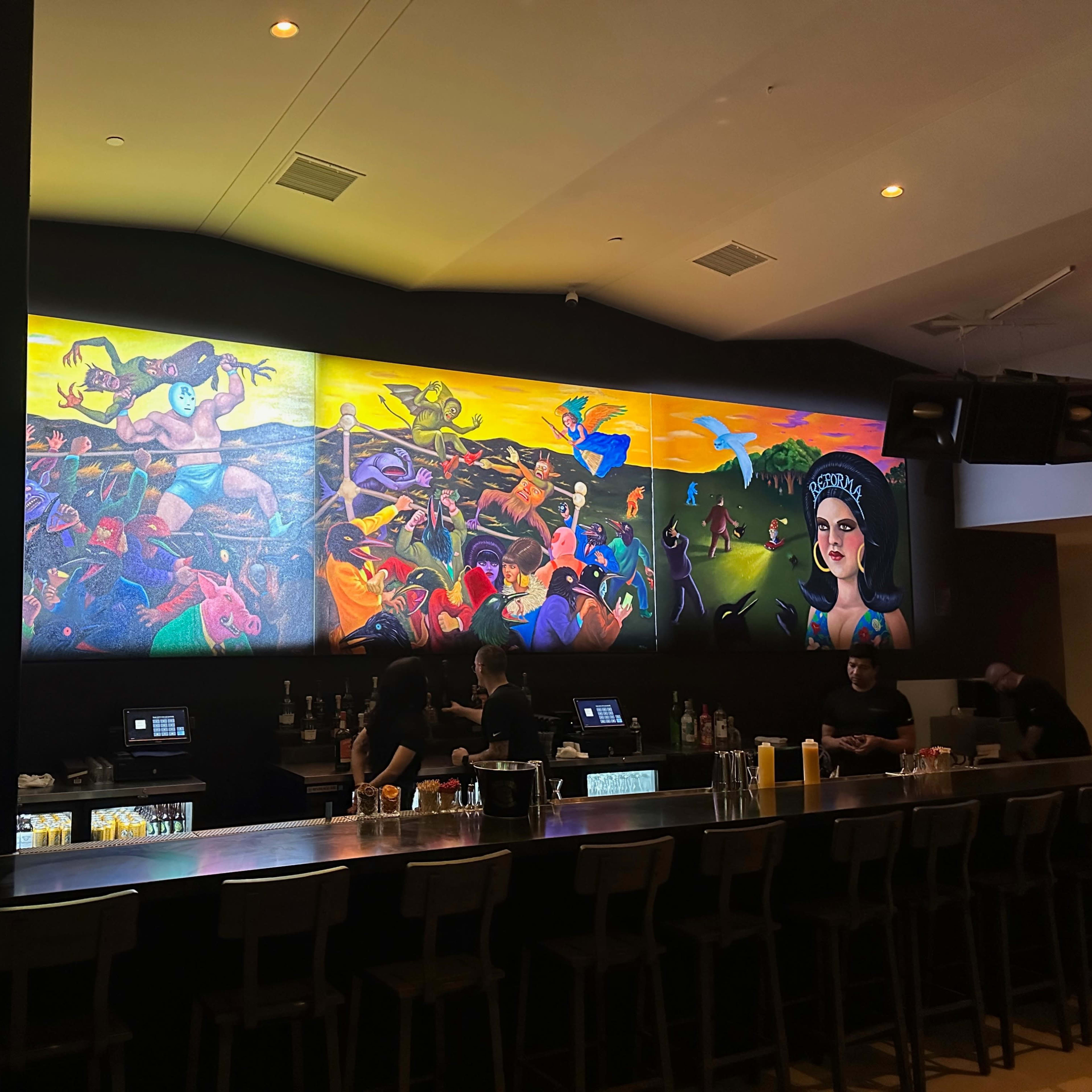 A vibrant mural on the wall of a bar, depicting various colorful figures and scenes, with bartenders working behind a long counter.