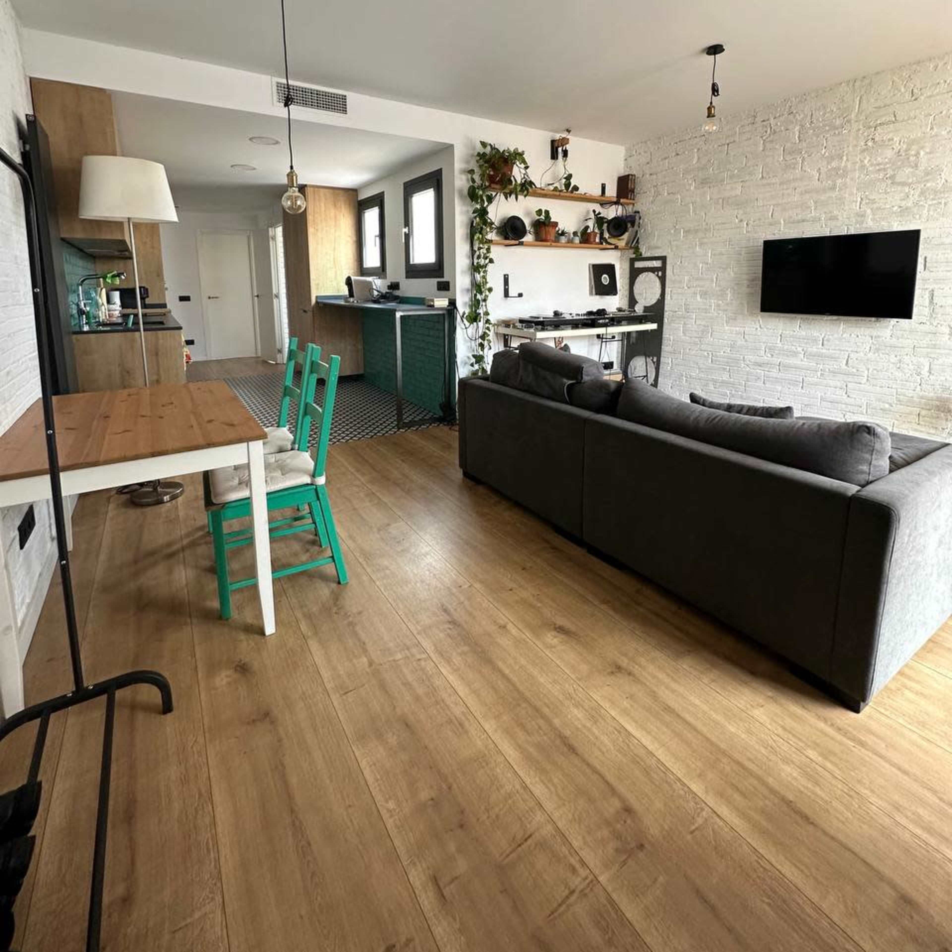 A modern living space featuring a gray sofa, a wooden dining table with green chairs, and a kitchen area with plants and shelves.