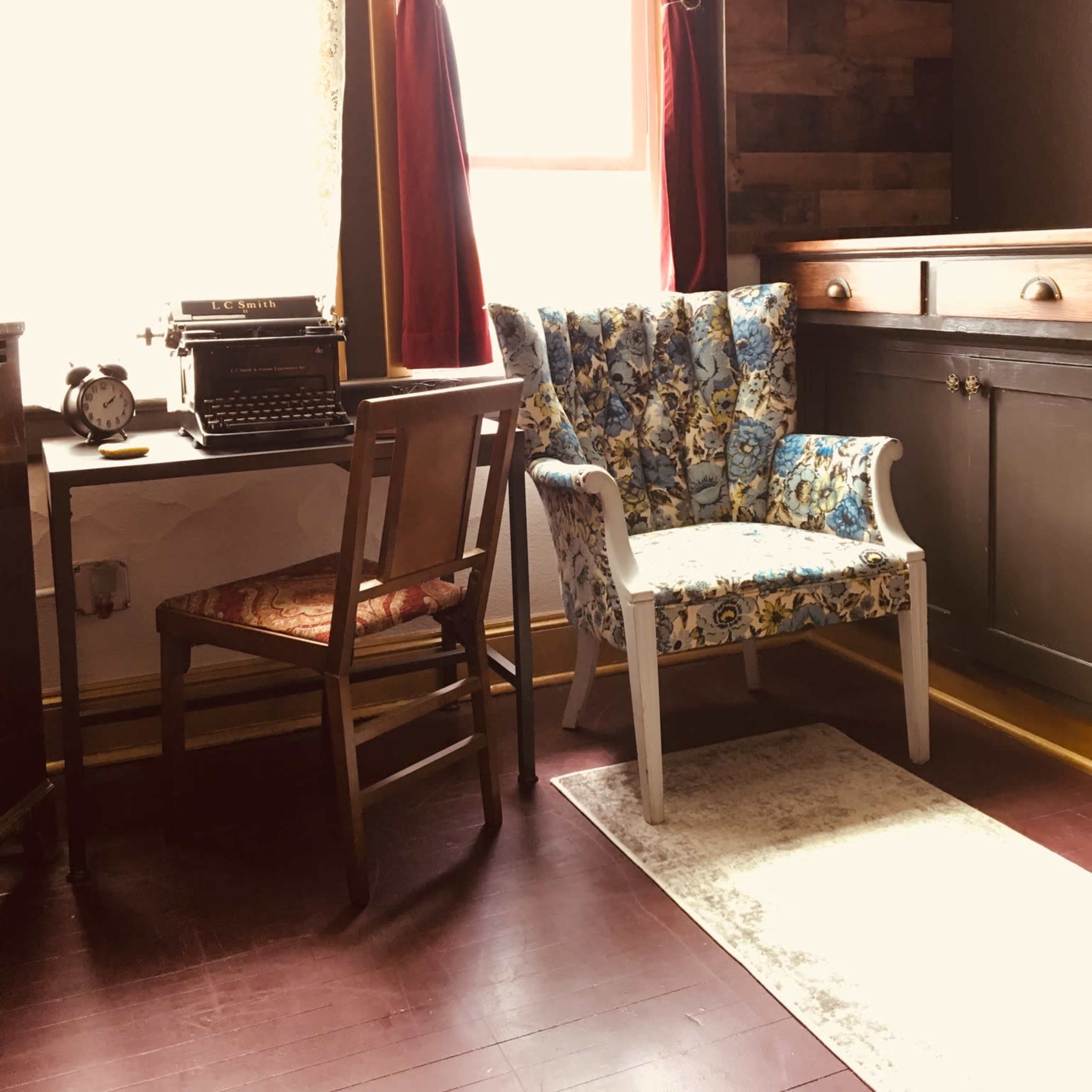 A vintage writing nook features a patterned armchair, a small desk with an antique typewriter, and an old clock, illuminated by natural light from a window.