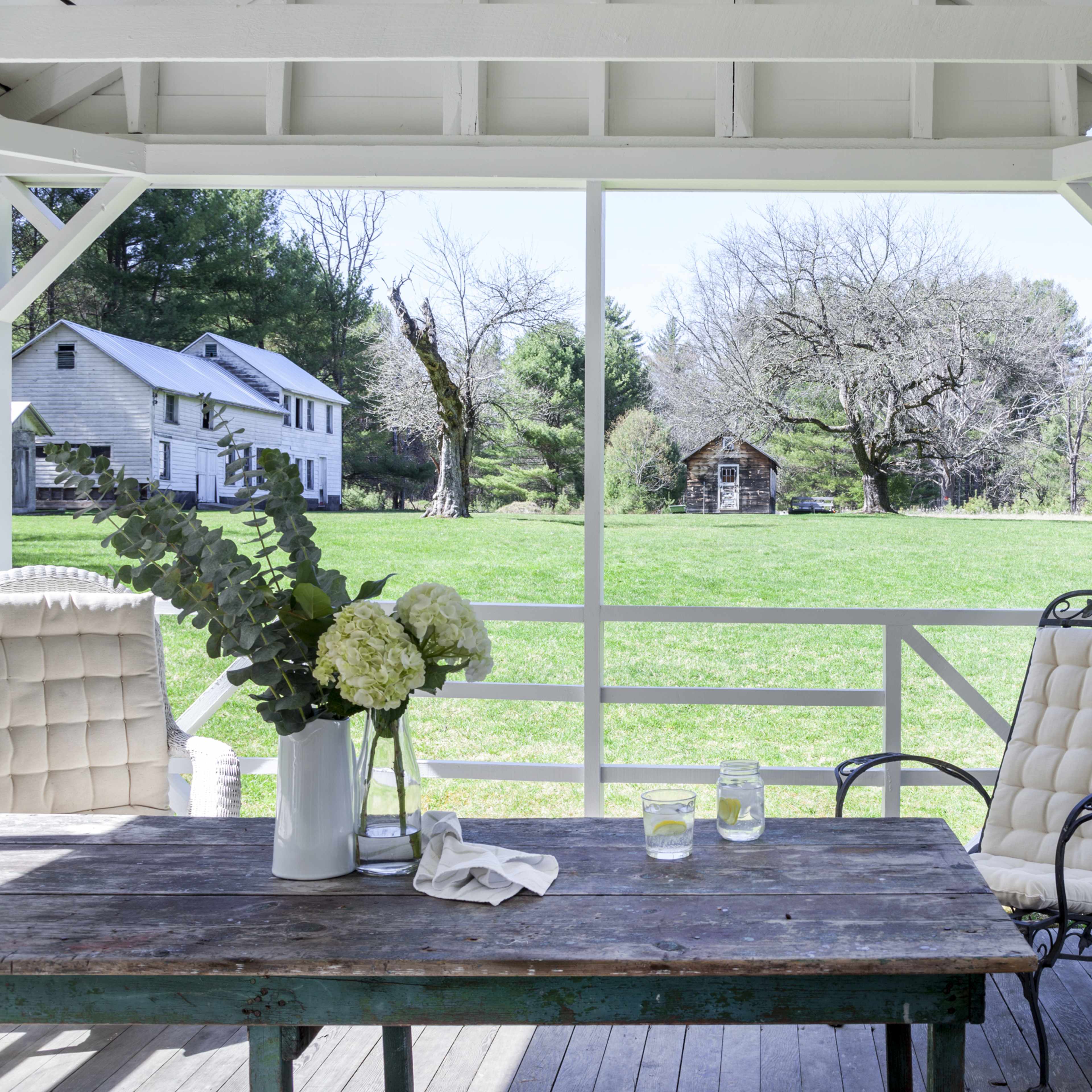 A rustic porch features a weathered wooden table with a vase of flowers and a metal chair, overlooking a grassy yard and a few distant structures.