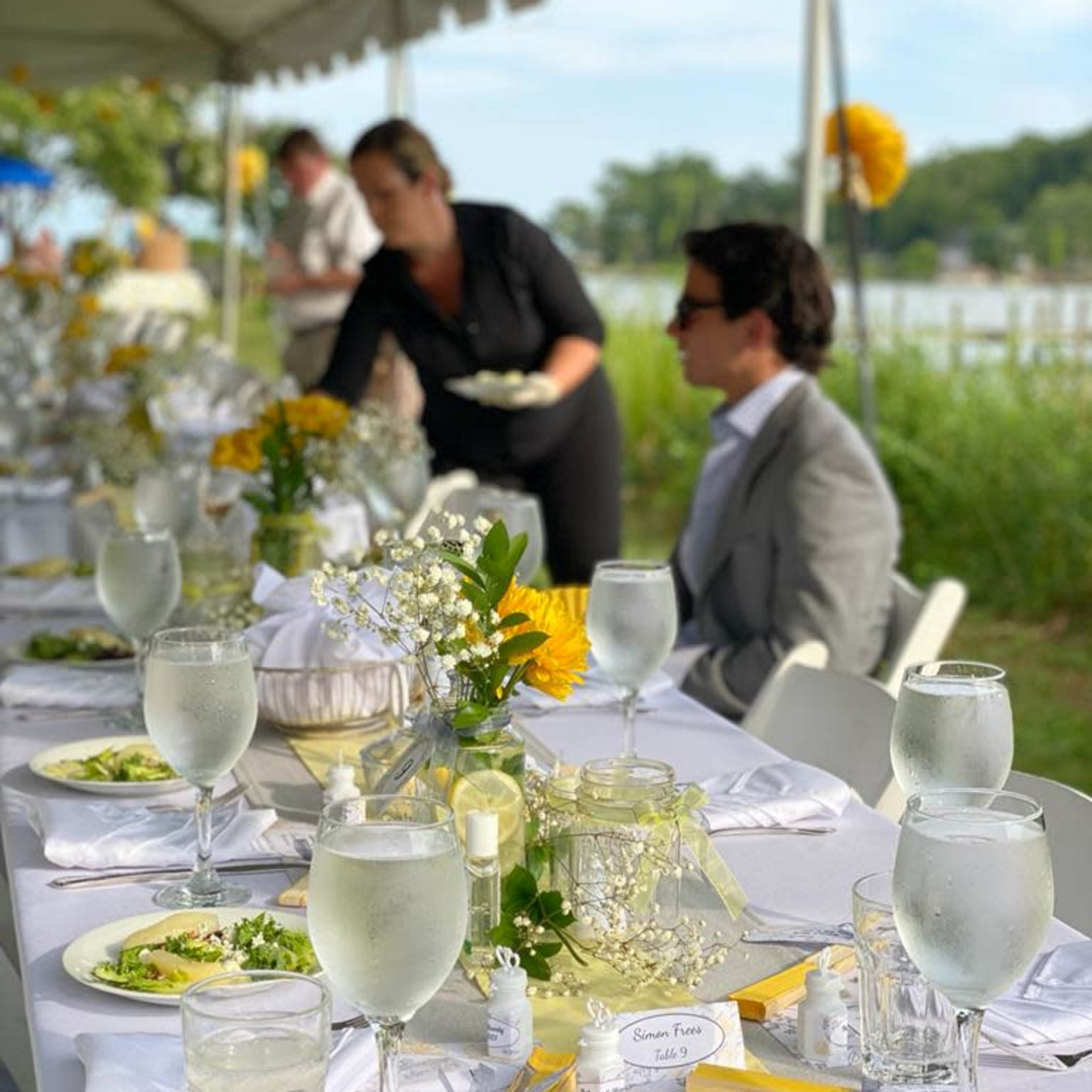 A long table elegantly set with glassware, floral decorations, and plates of salad occupies an outdoor space near a body of water, with a server in the background.