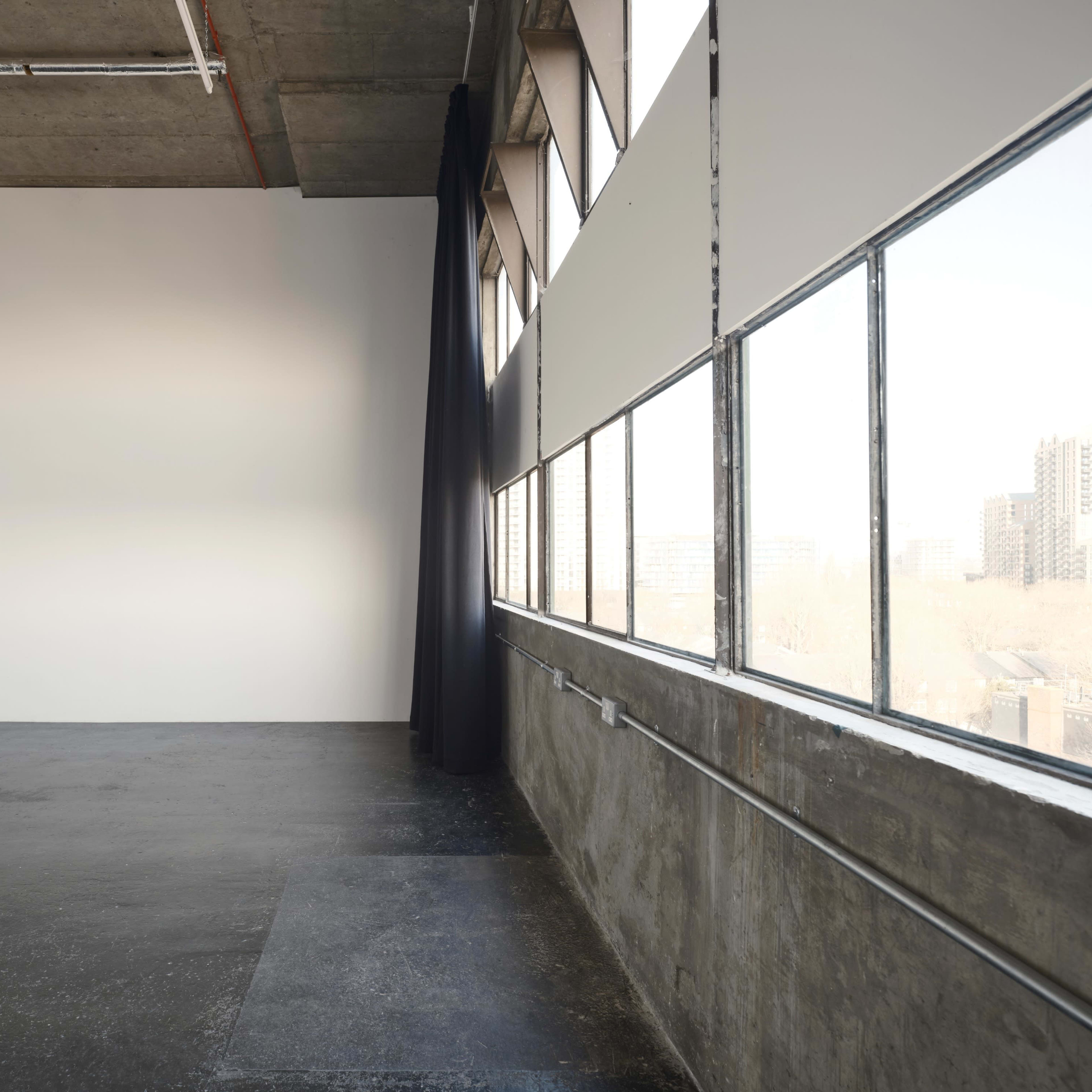 A spacious, industrial-style room with large windows, concrete walls, and a dark curtain partially drawn.