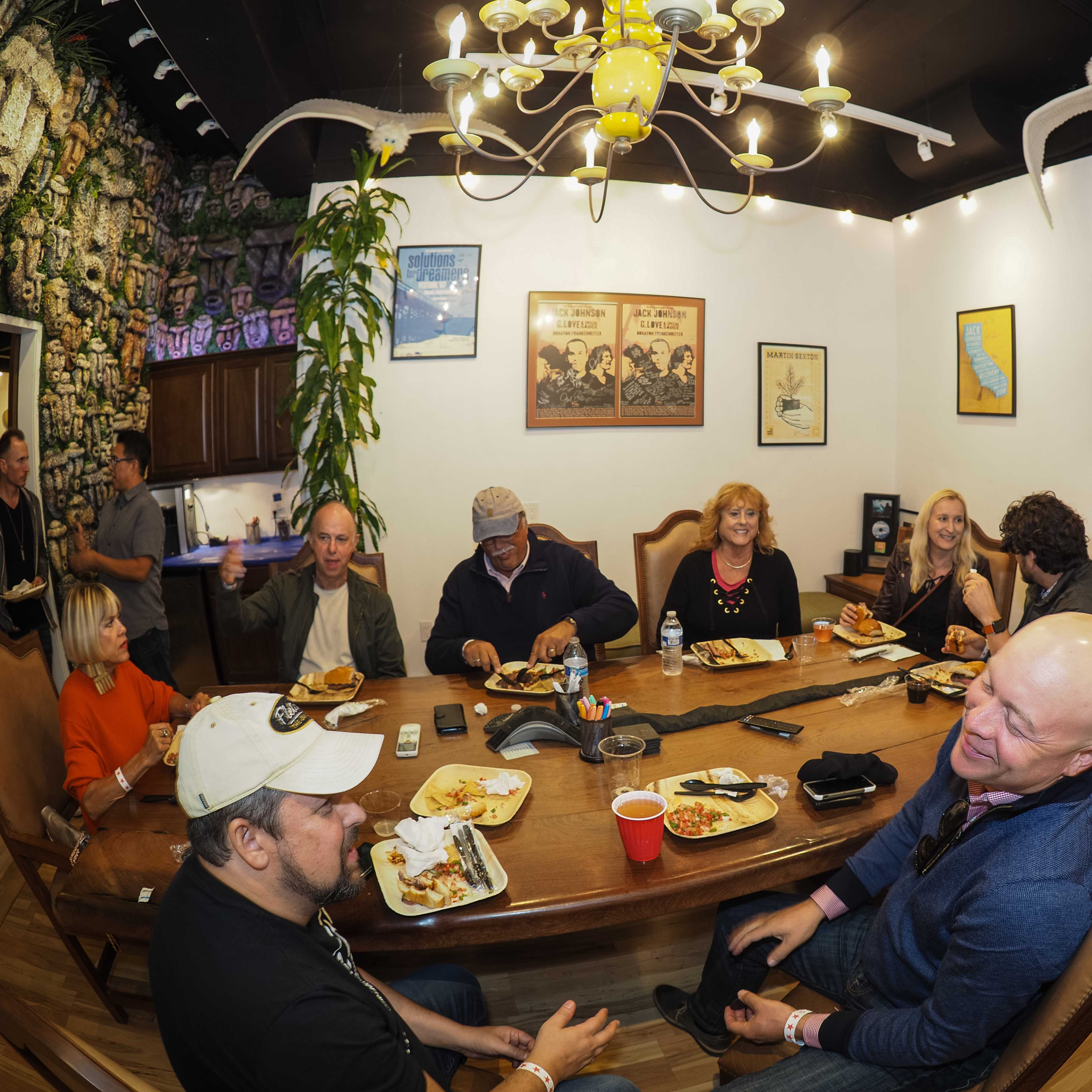 A group of people gathers around a large table, enjoying food and drinks while engaged in conversation in a brightly lit room decorated with various pictures and plants.