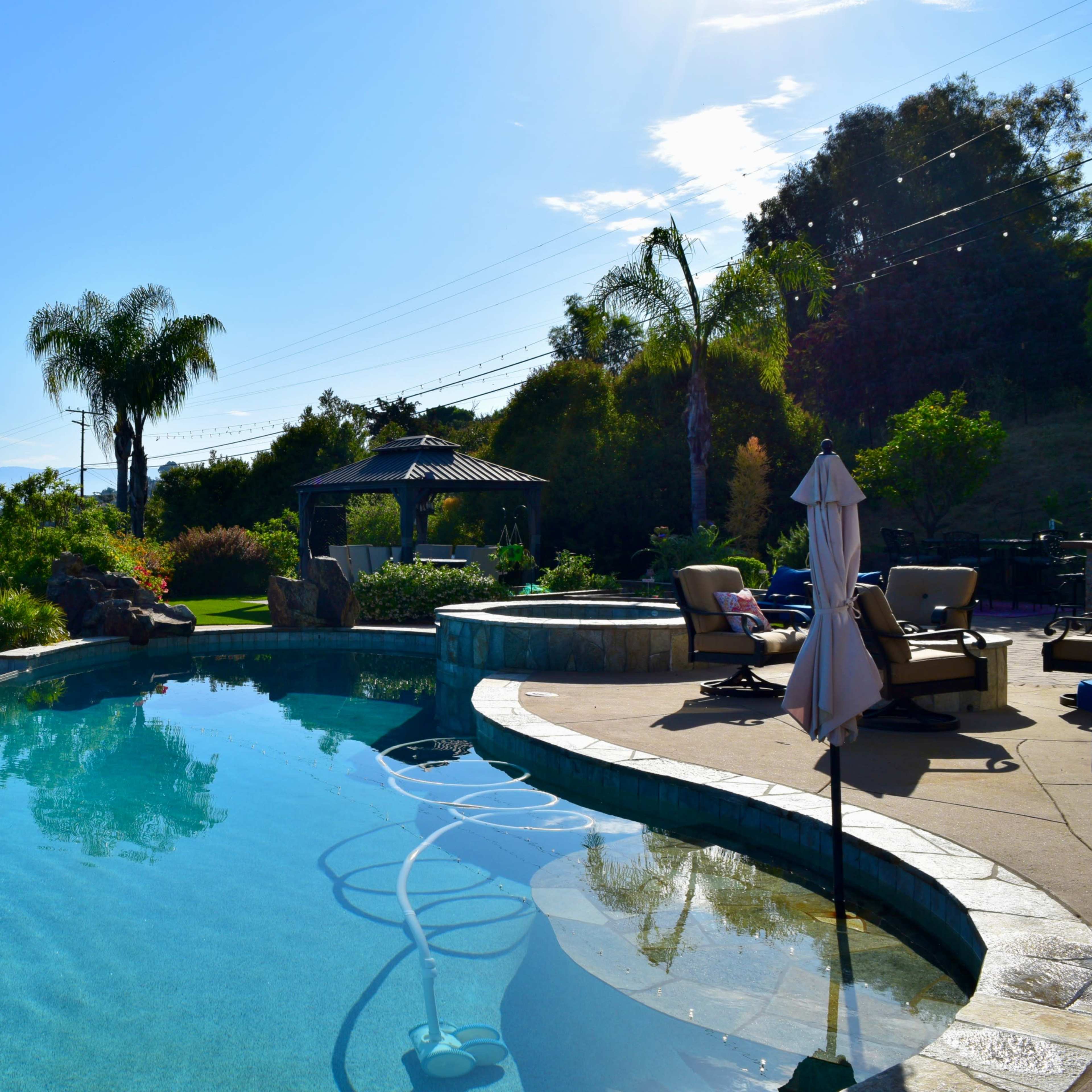 A clear swimming pool surrounded by lush greenery and a gazebo in the background, set against a mountainous landscape under a blue sky.