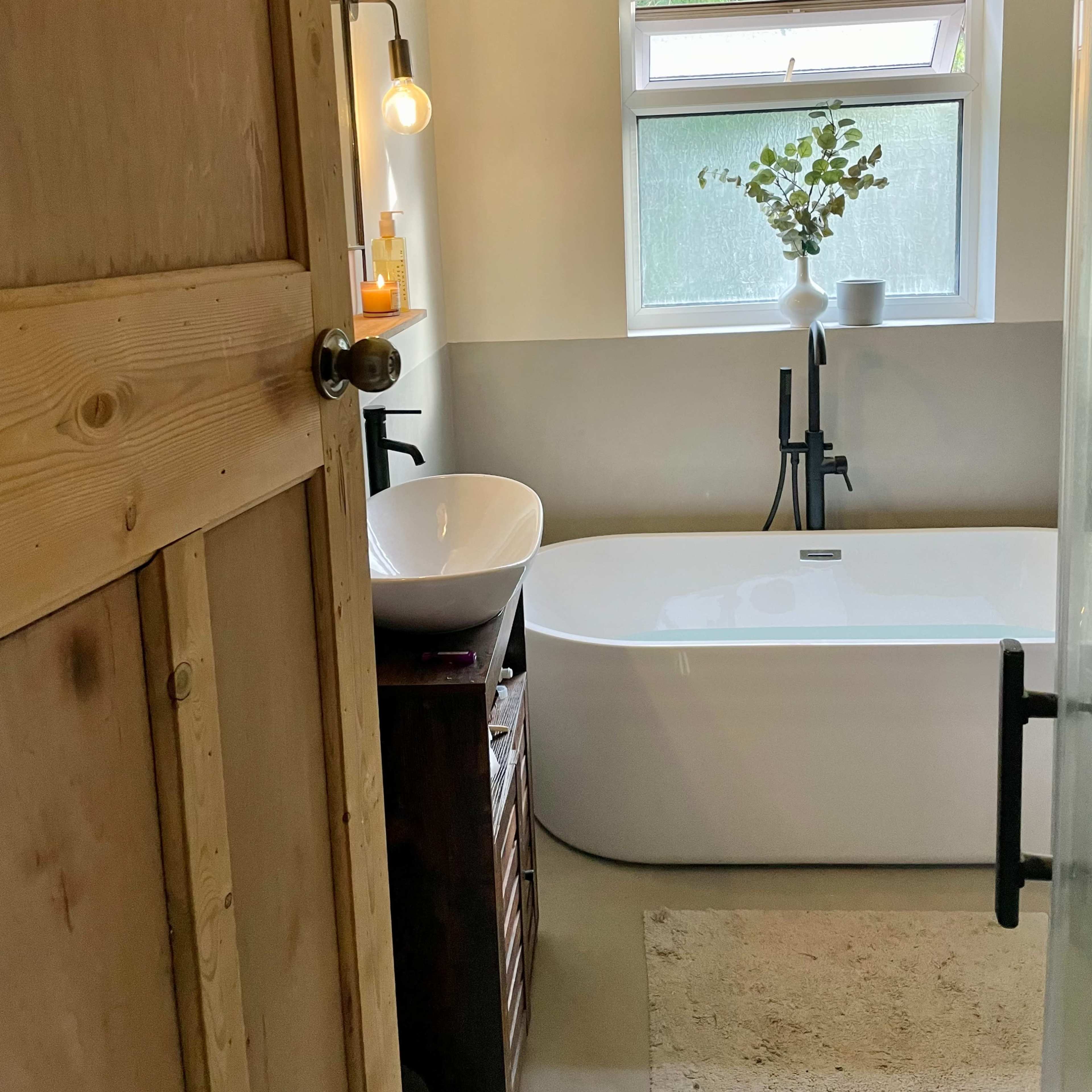 A modern bathroom featuring a freestanding bathtub, a sink with a wall-mounted faucet, and a window allowing natural light, along with a wooden door and minimal decor.