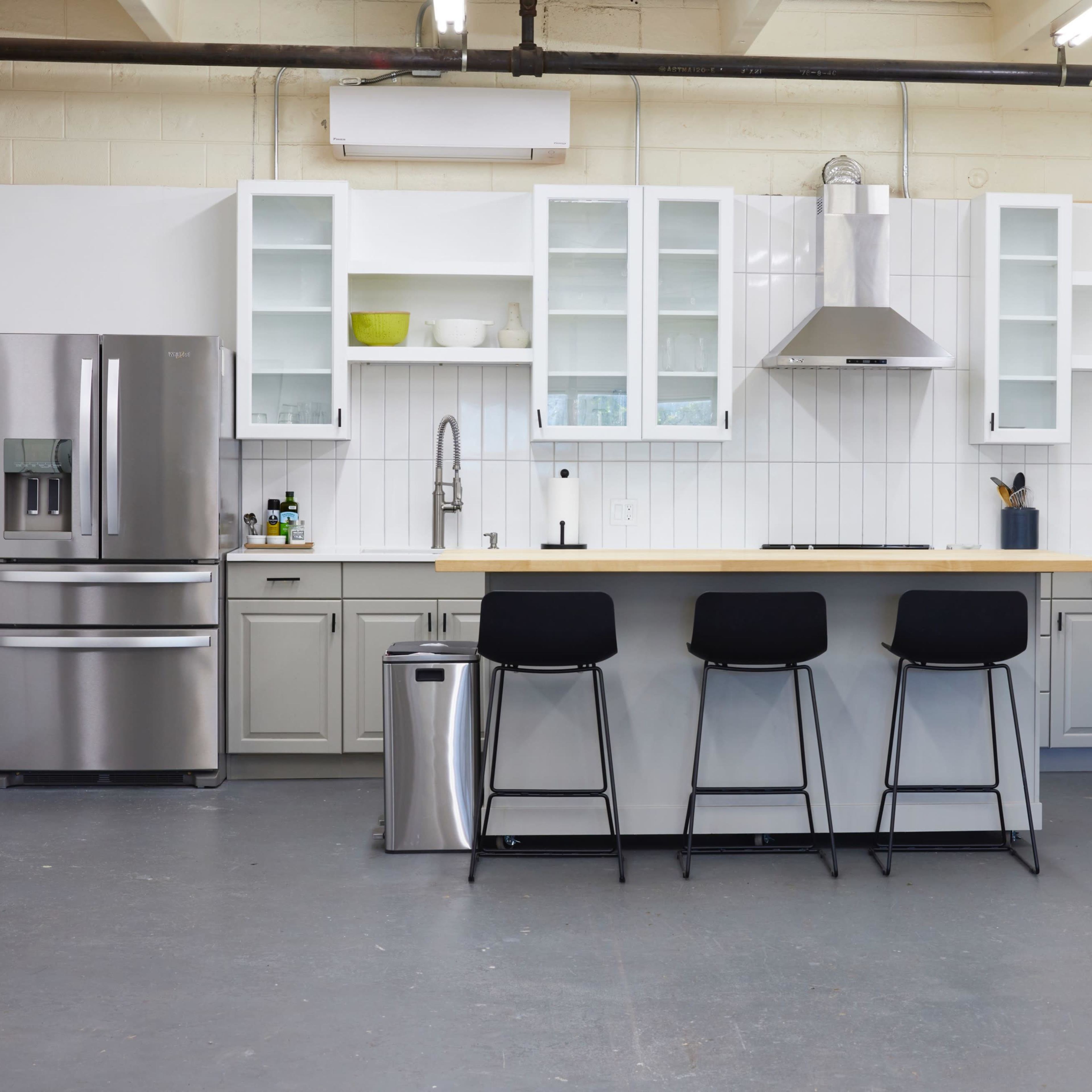 A modern kitchen featuring a central island with three black stools, stainless steel appliances, and white cabinetry against a gray floor.