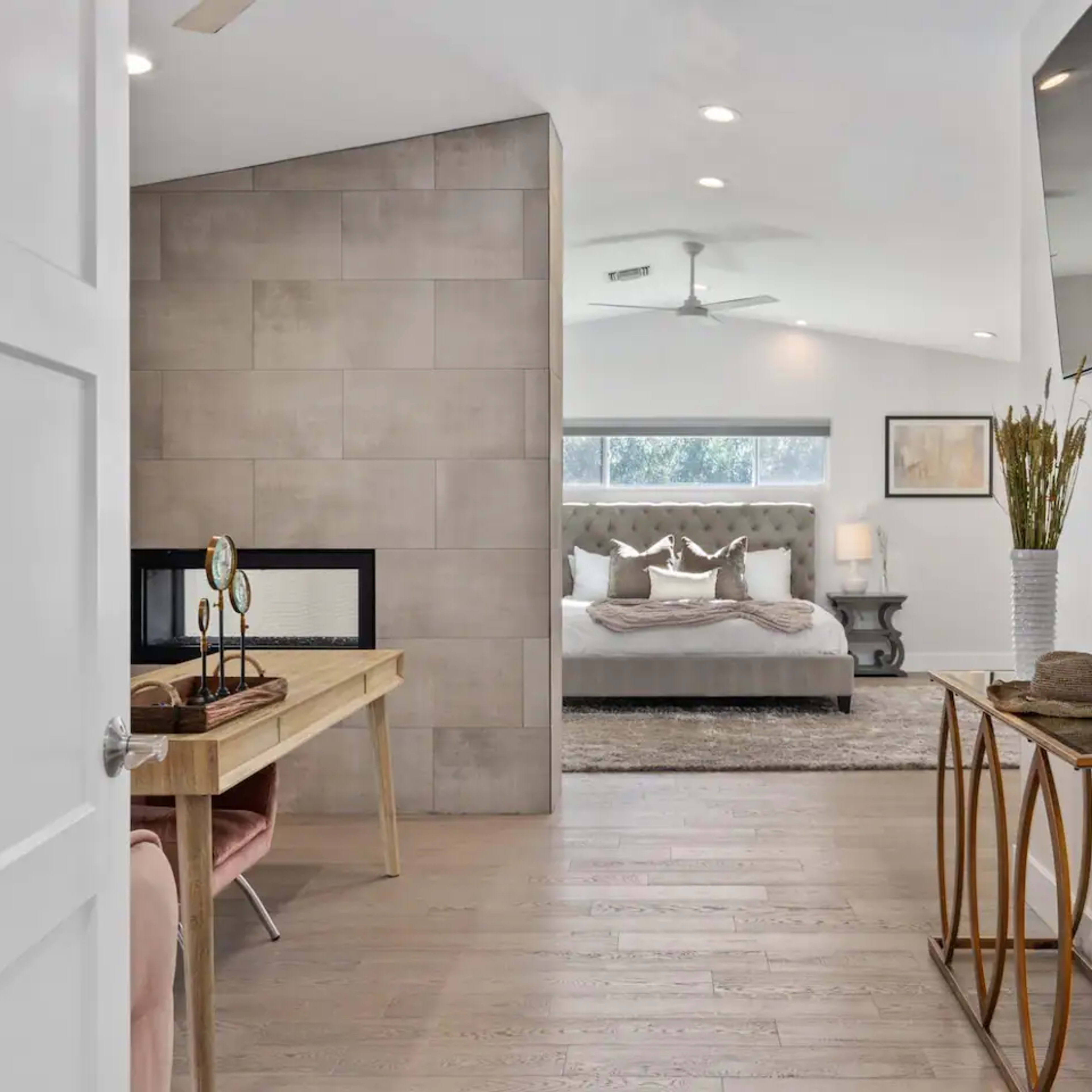 A modern, well-lit bedroom is visible through an open doorway, featuring a bed with neutral bedding, a decorative area rug, and a cozy seating area.