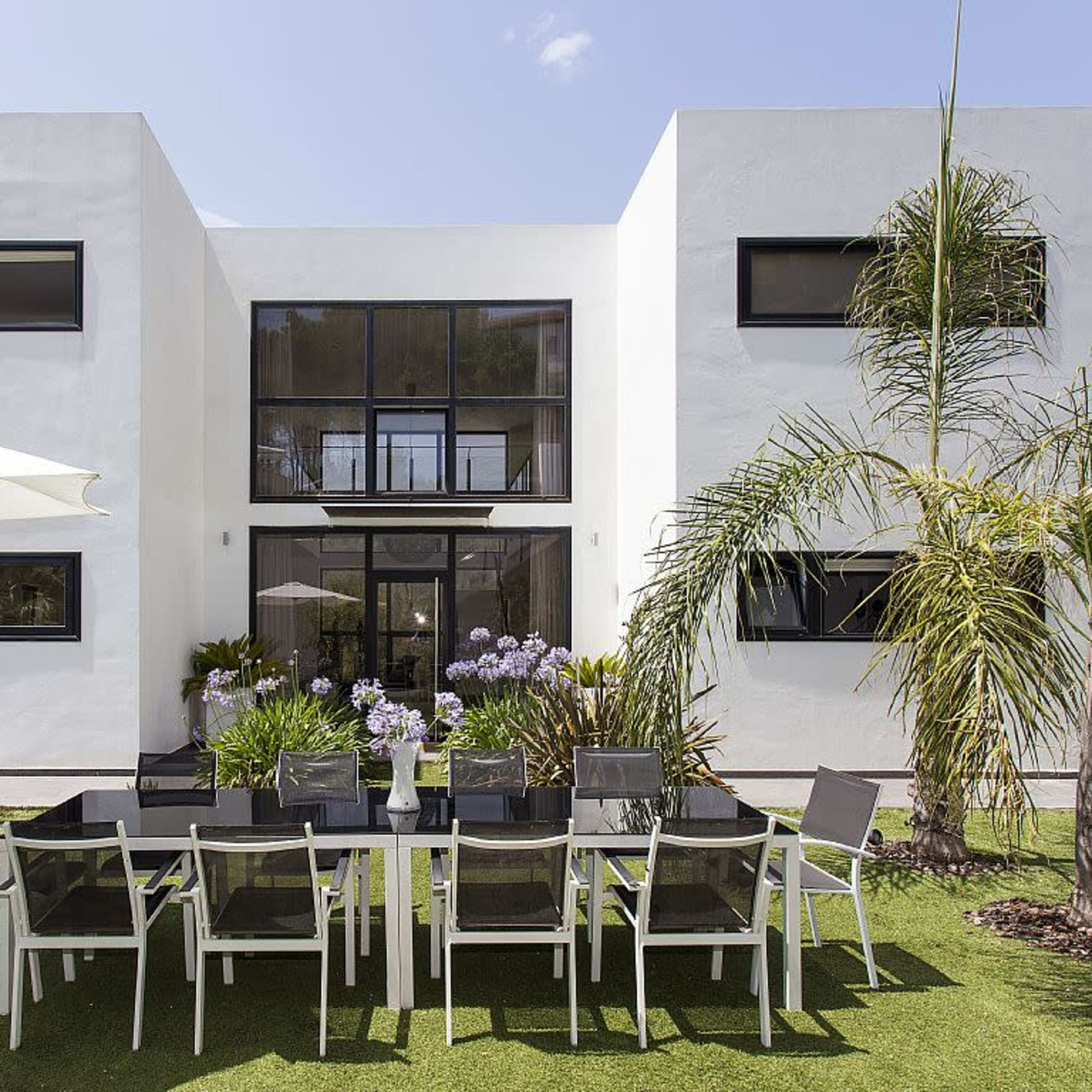 A modern two-story house with large windows features a spacious outdoor dining area set with a long table and chairs on a grassy lawn.