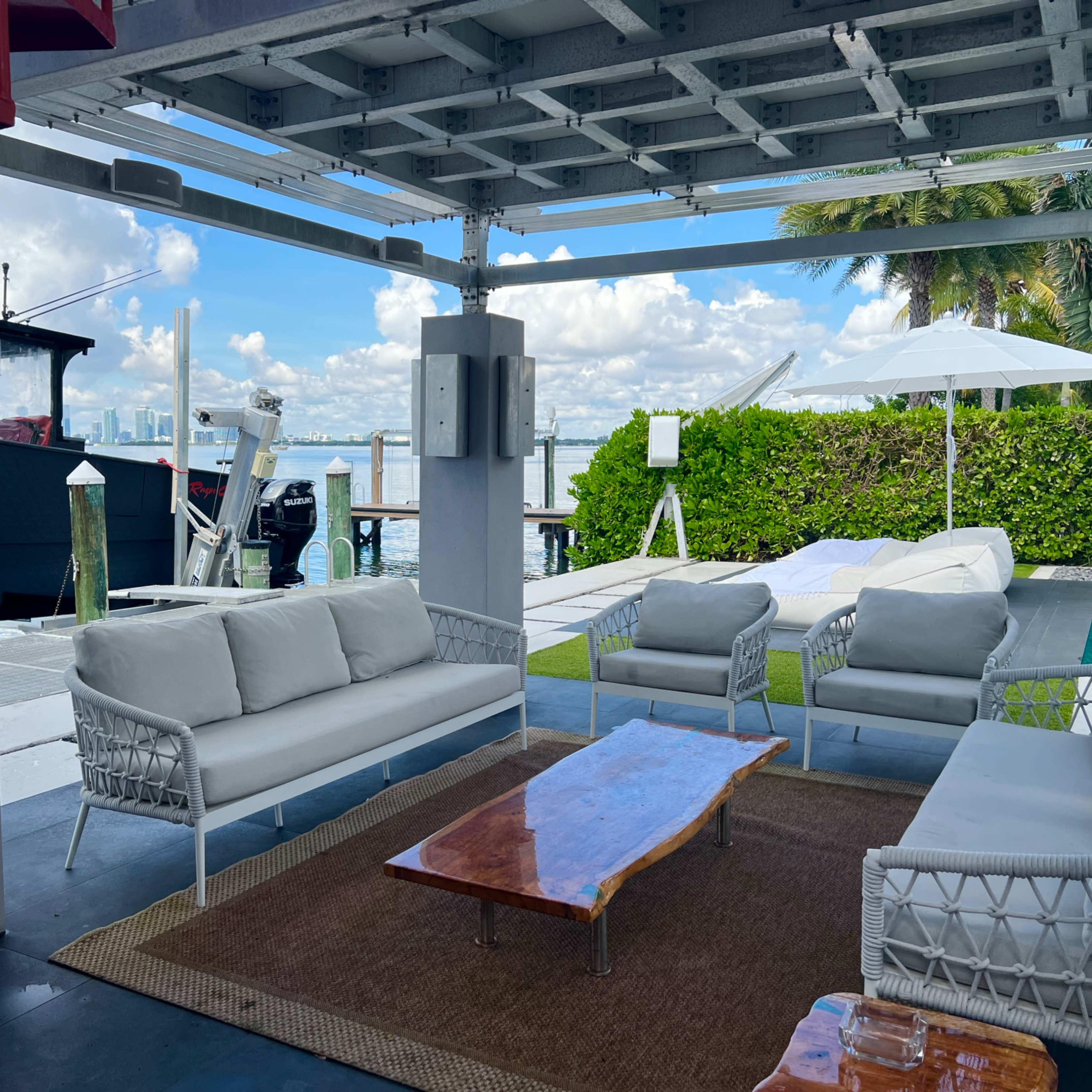 A waterfront patio features white wicker furniture arranged around a wooden coffee table, with a view of a marina and boats in the background.