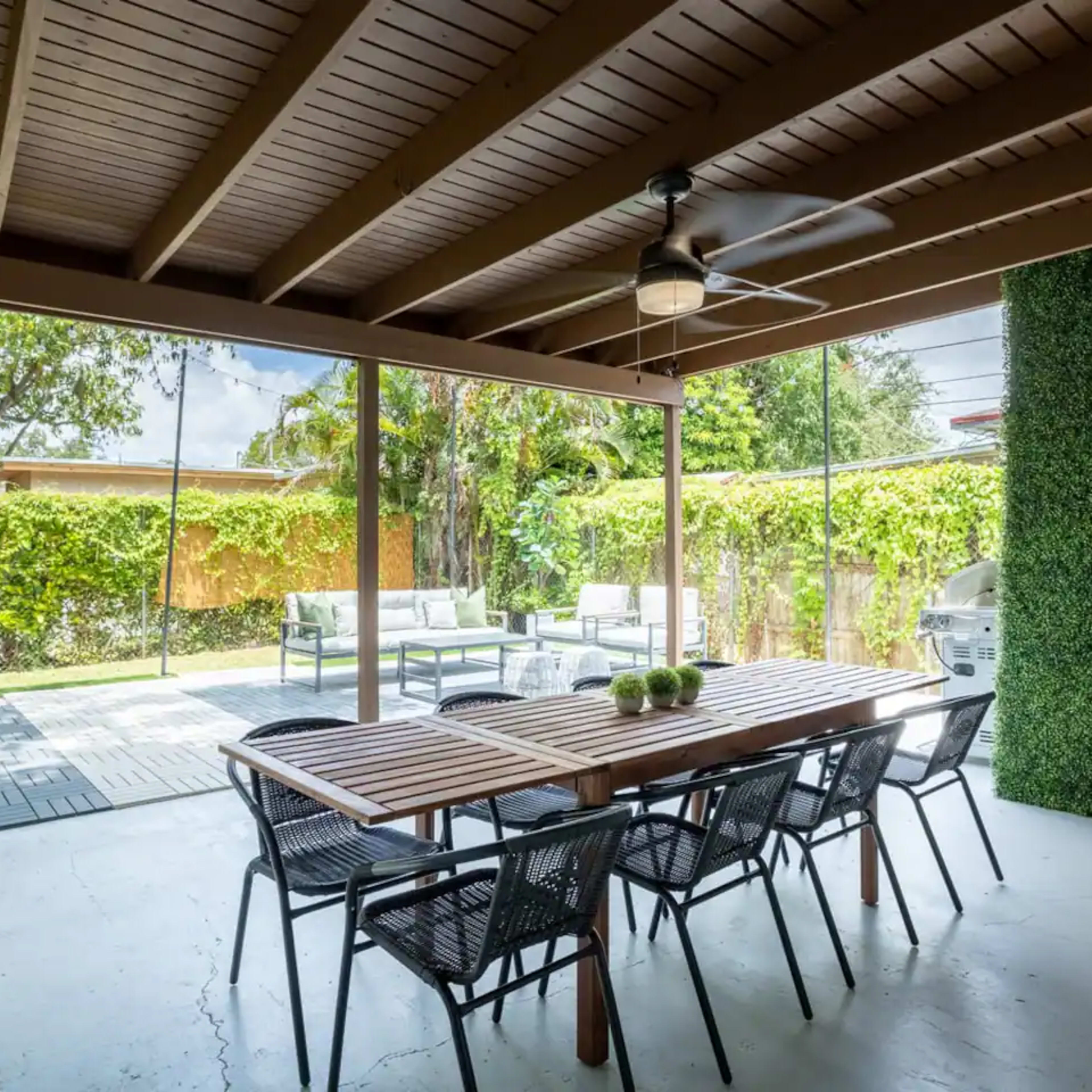 A sheltered outdoor patio with a wooden dining table, black chairs, and a view of a green space beyond.