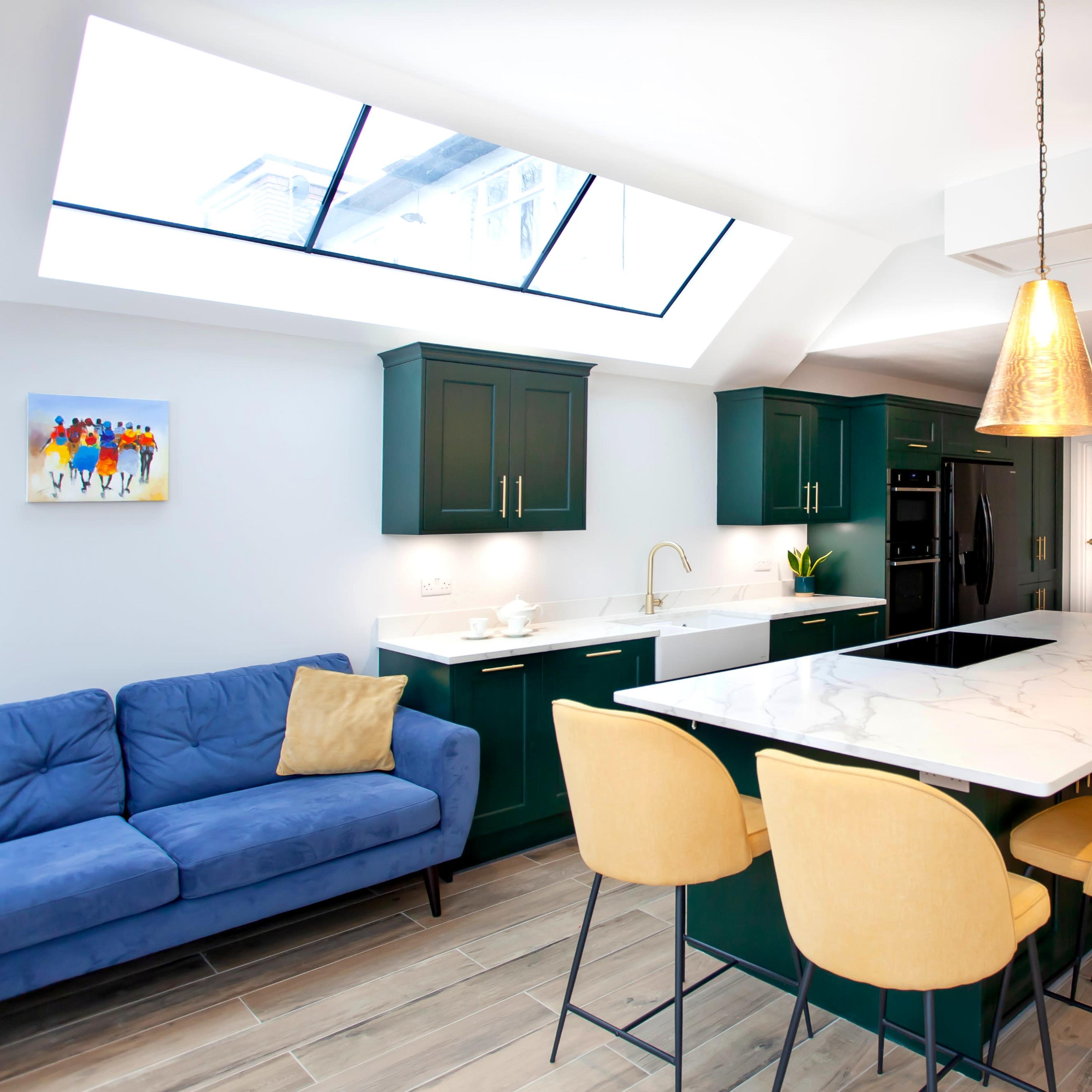 The kitchen features green cabinetry, a large marble island with yellow chairs, and a skylight above.