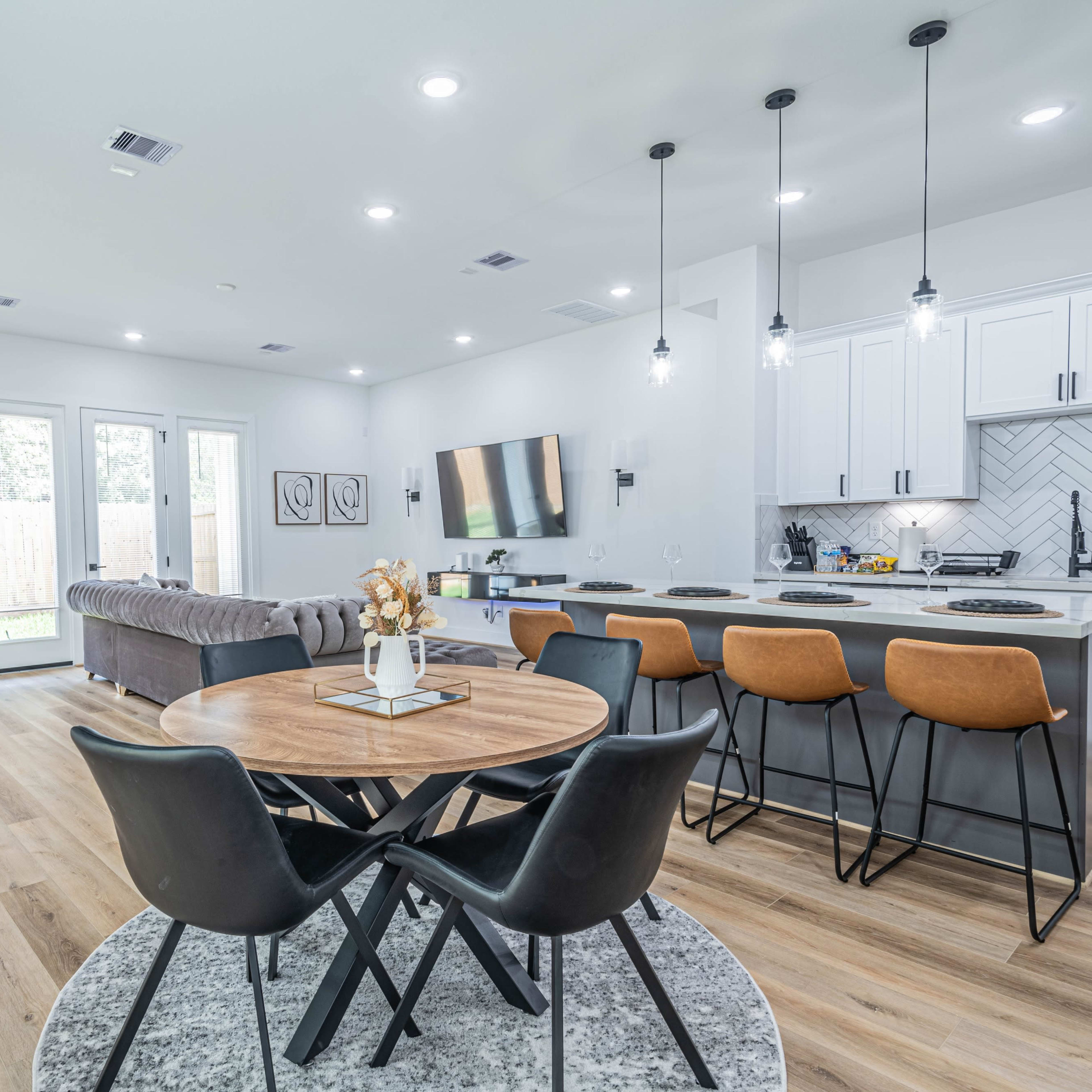 A modern living space featuring a dining area with a round table and black chairs, a kitchen with white cabinetry, and a cozy seating area with a large couch and a television.