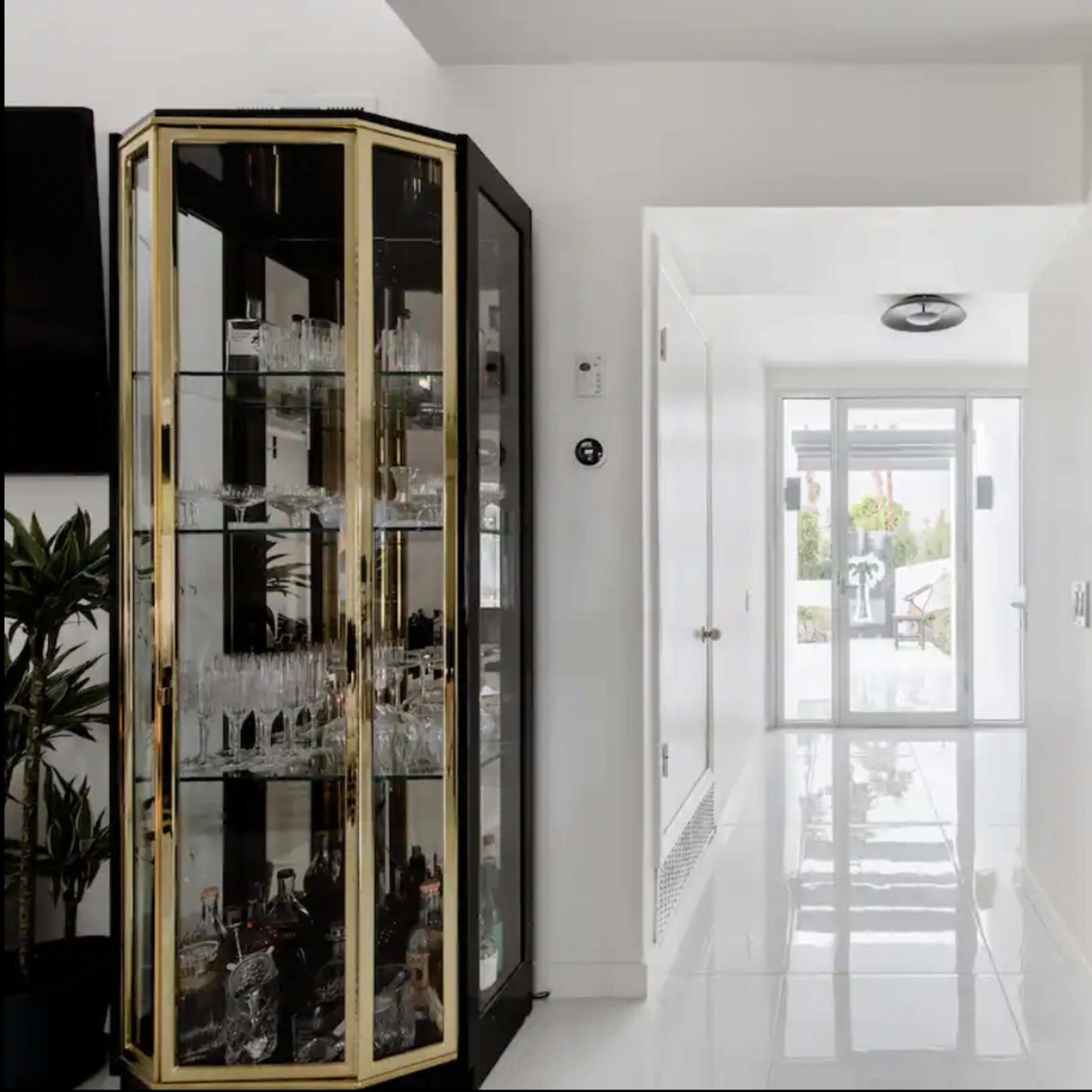 A modern interior with a glass display cabinet on the left and a stainless steel refrigerator on the right, leading to a bright, open hallway.