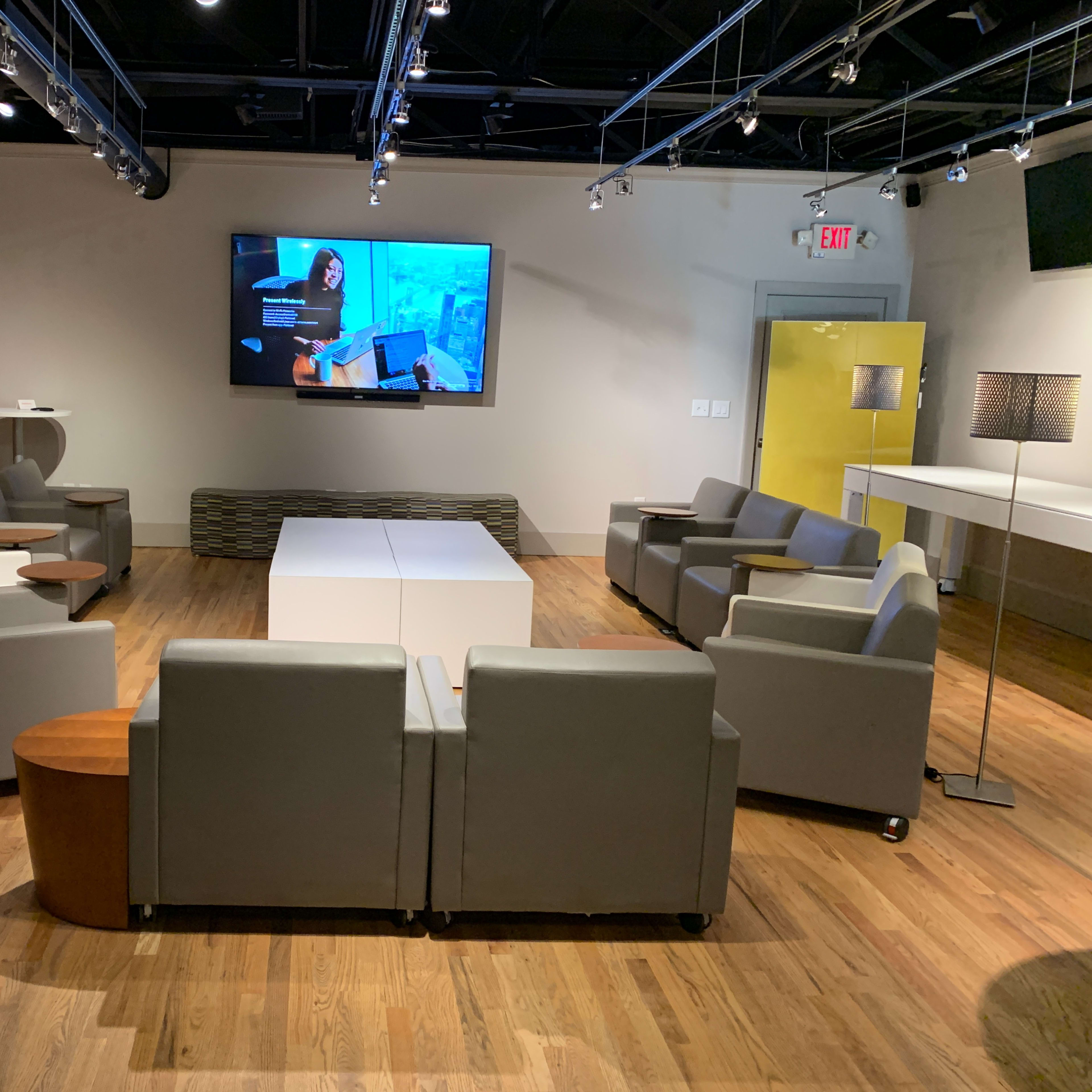 A modern lounge area with gray seating, a large screen on the wall, and wooden flooring.