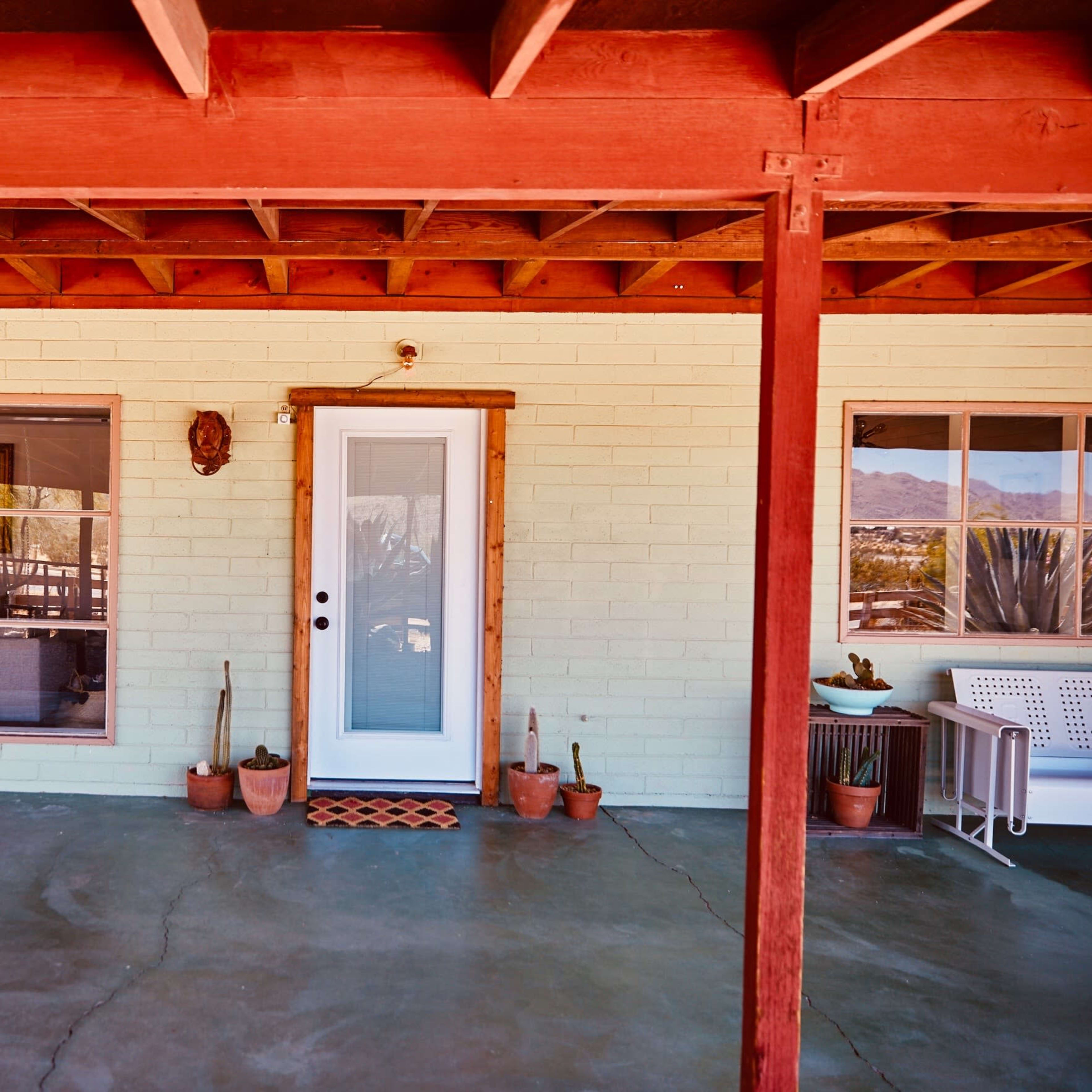 A front porch area of a building with a light-colored brick wall, a central door, large windows on one side, and potted plants.